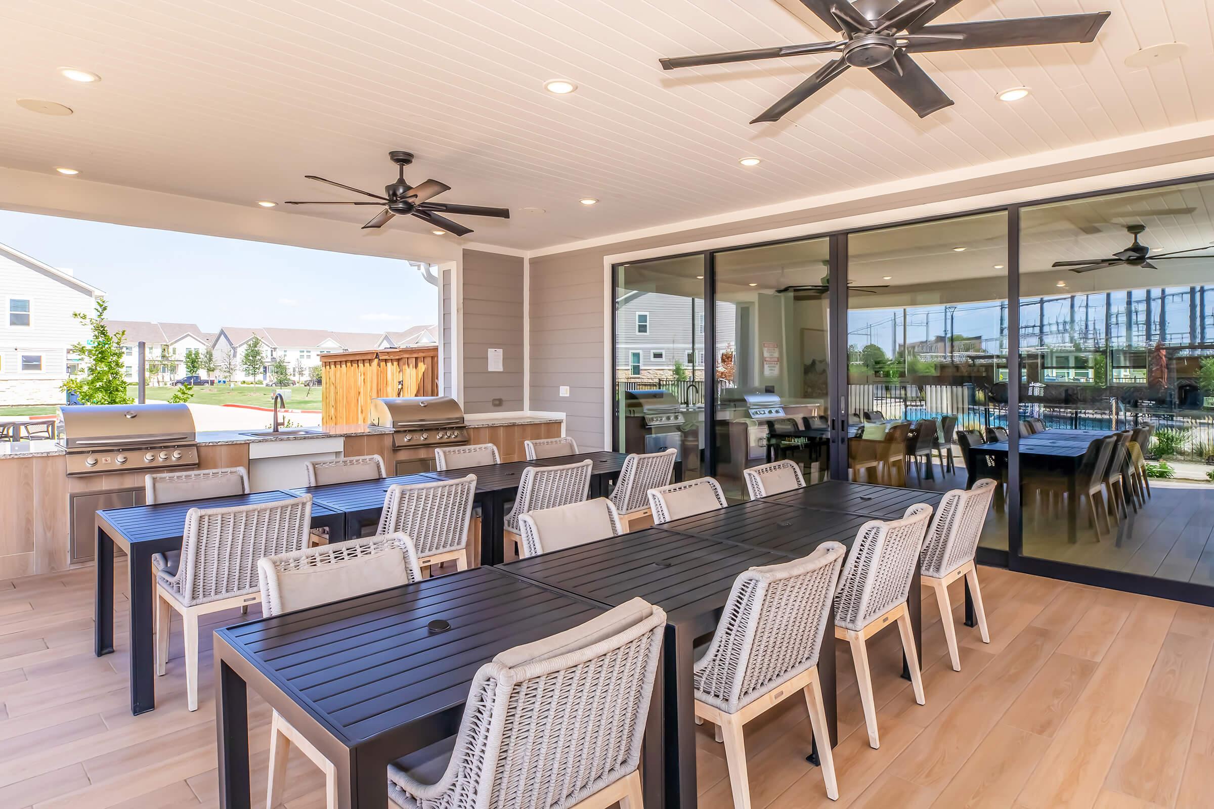 A spacious outdoor patio area featuring a long black dining table surrounded by light-colored chairs. There are two ceiling fans overhead and a grilling station with stainless steel grills. Large glass doors open to a view of a well-manicured lawn and nearby homes, creating an inviting atmosphere for gatherings.