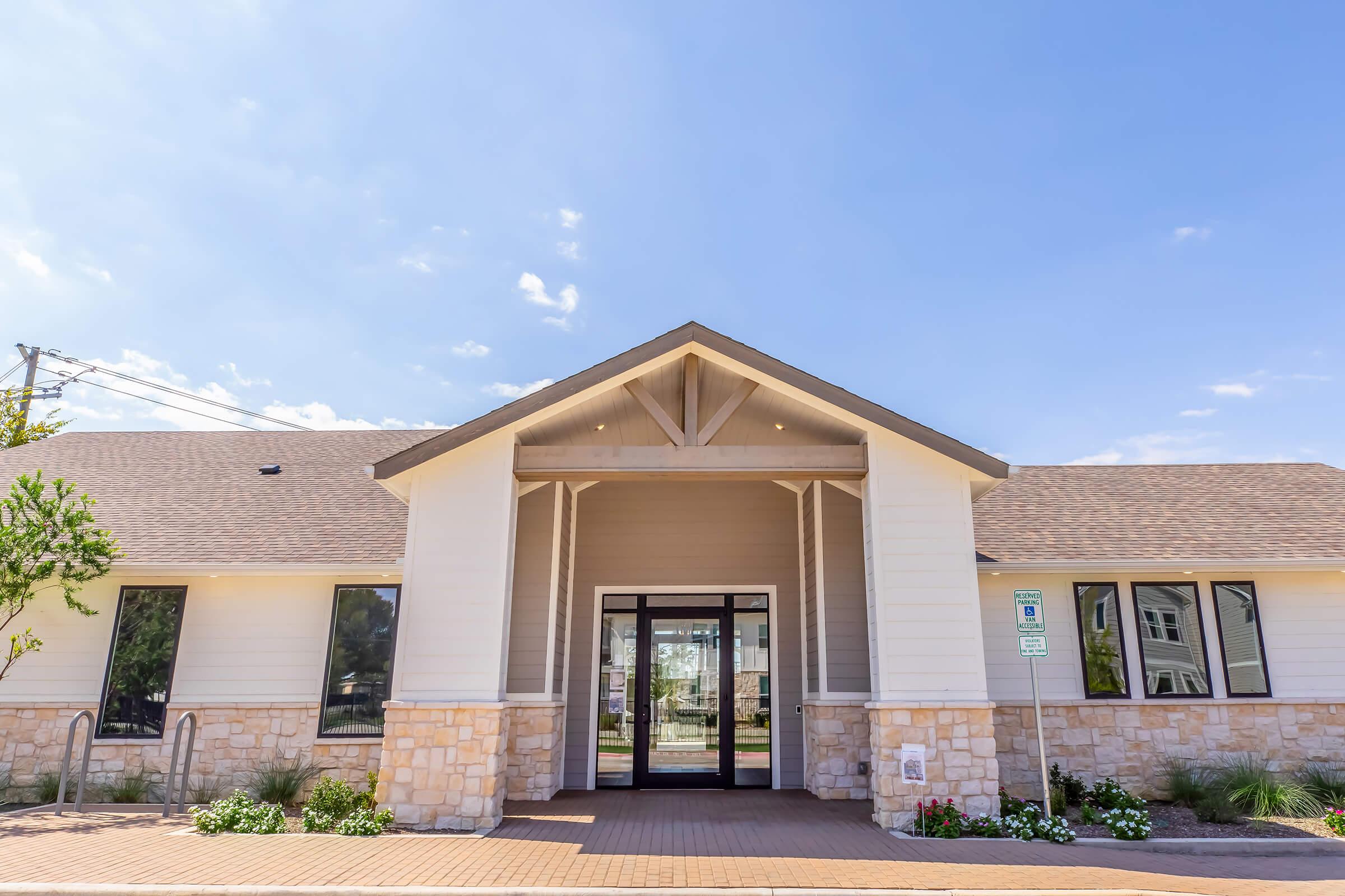 A modern building with a stone and wooden facade, featuring a large entryway with glass doors. The roof is peaked, and there are decorative elements around the entrance. In front, there are neatly landscaped flower beds and a clear blue sky above. The pathway leading to the entrance is paved.
