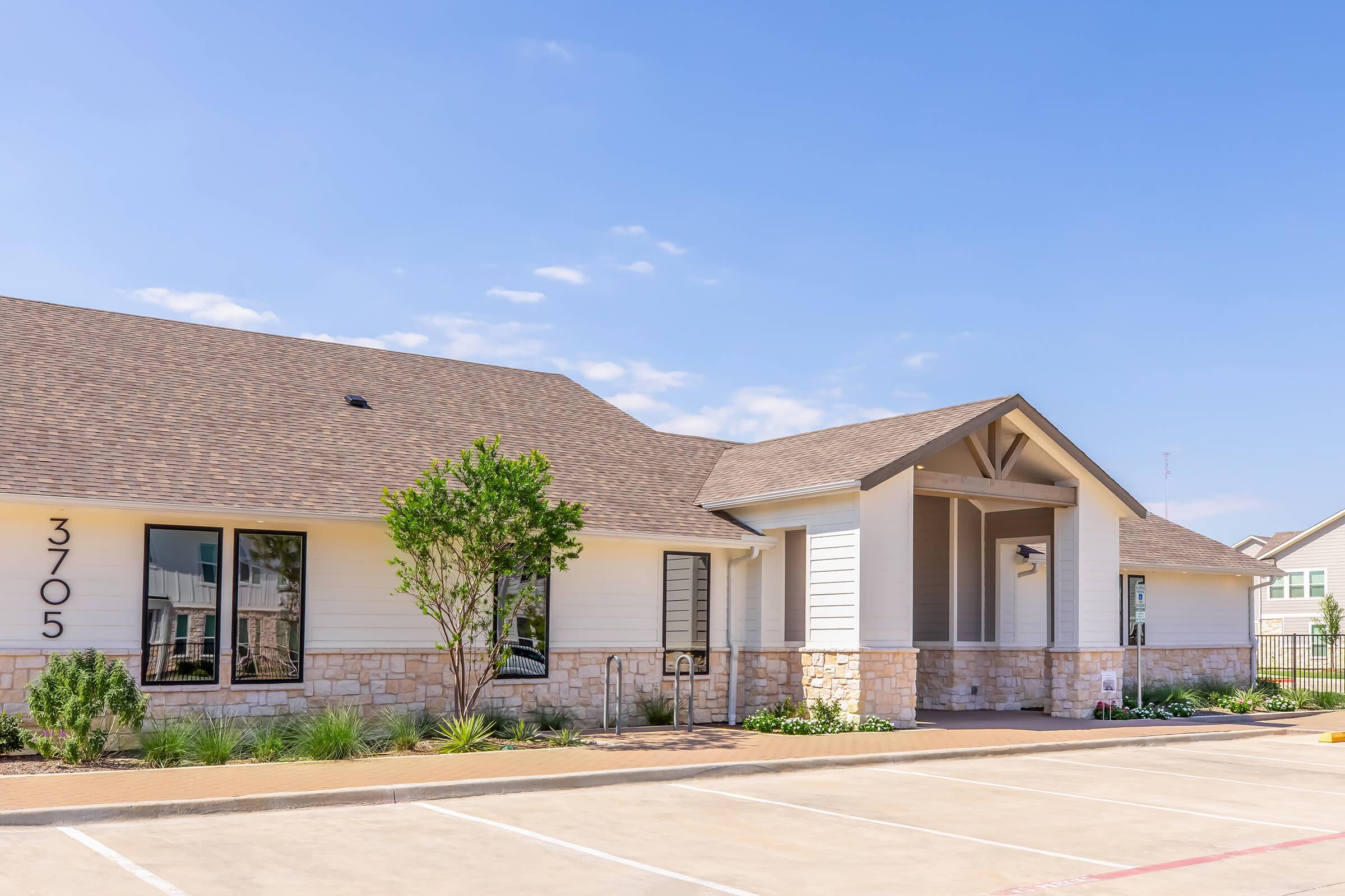 A modern building with a light-colored exterior featuring stone accents, large windows, and a sloped roof. The entrance has a covered porch supported by wooden beams. Surrounding the building are landscaped areas with shrubs and small plants. The sky is clear with a few clouds.