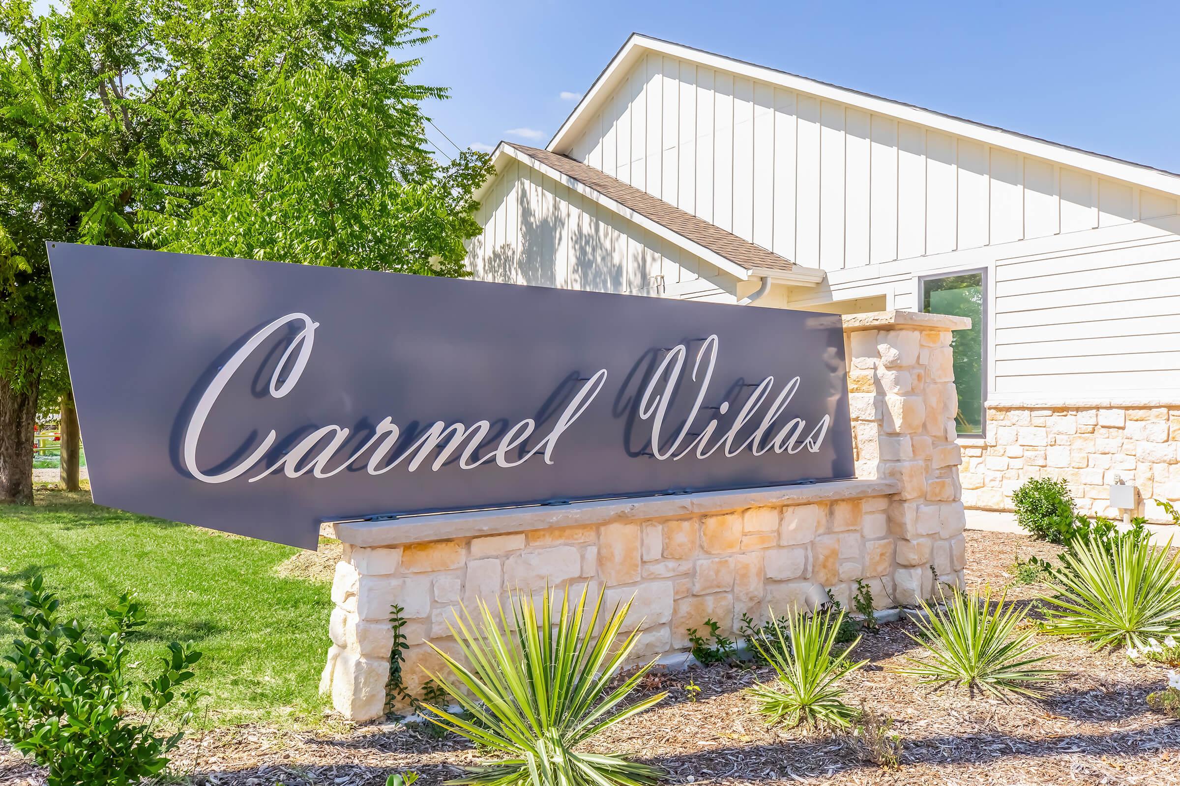Signage for Carmel Villas displayed prominently in front of a building, with a modern design featuring elegant cursive lettering. The sign is mounted on a stone base, surrounded by landscaped greenery and native plants under a clear blue sky.