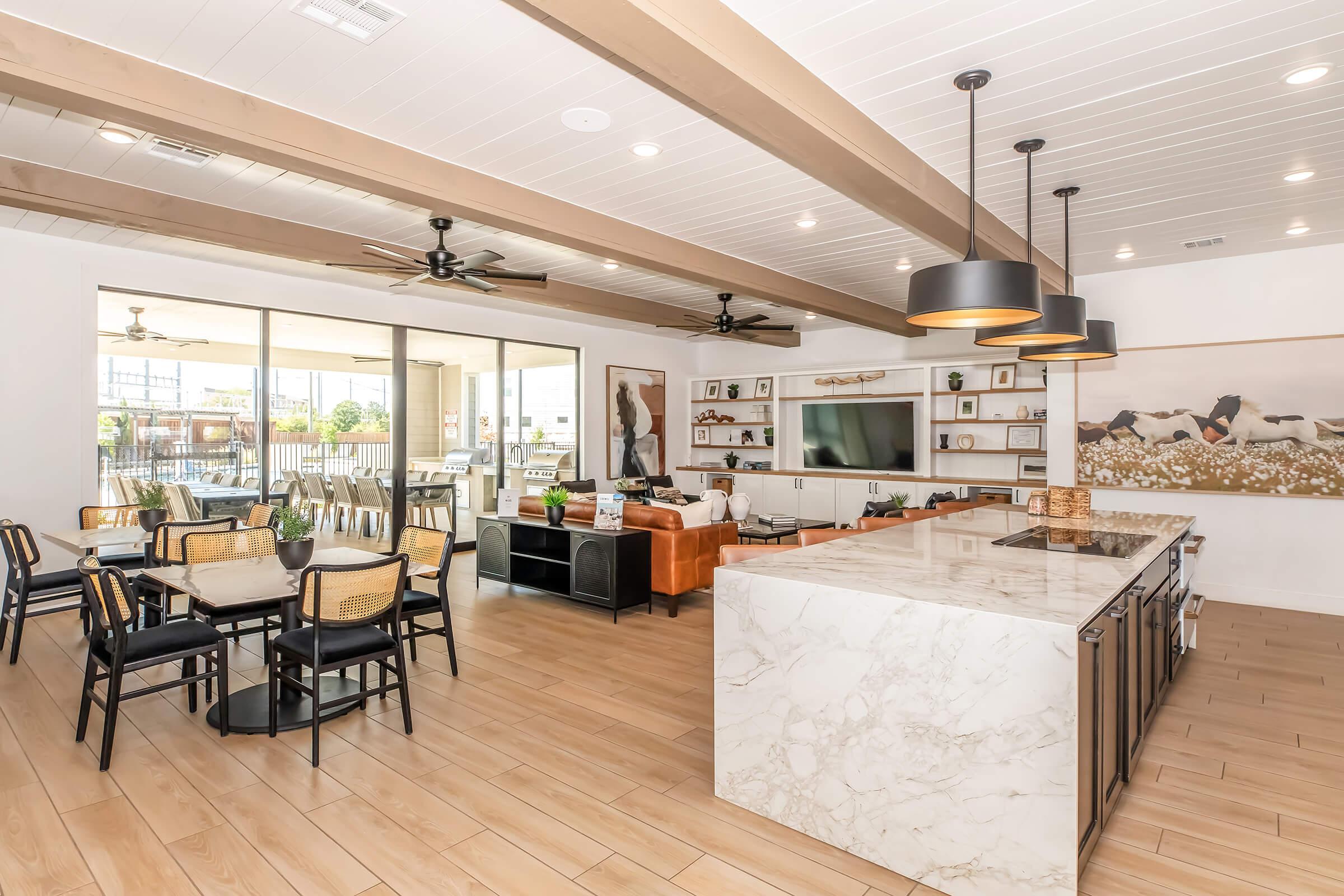 A spacious modern living area featuring a kitchen island with marble countertops, a dining table, and a comfortable seating area. Large sliding glass doors lead to an outdoor space, and stylish ceiling fans enhance the open ambiance. Decorative elements include artwork and shelving with various items.