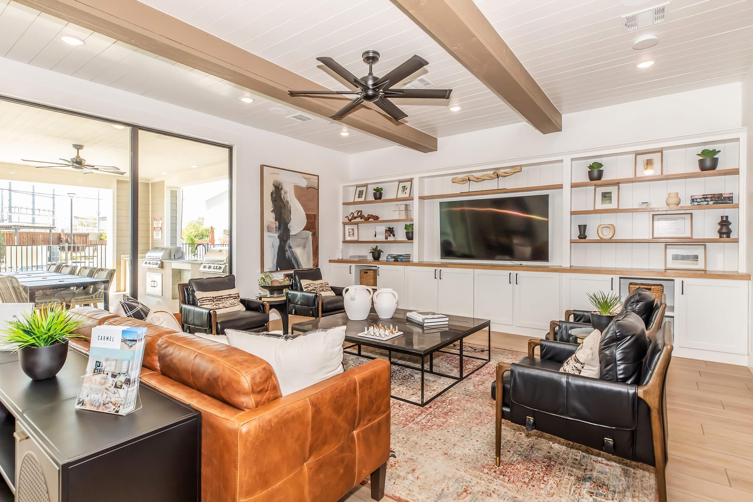 A modern living room featuring a brown leather sofa, black chairs, and a coffee table. There are decorative plants, artwork on the walls, and a large flat-screen TV mounted on a white cabinet. Natural light streams in through large sliding glass doors, revealing an outdoor area.