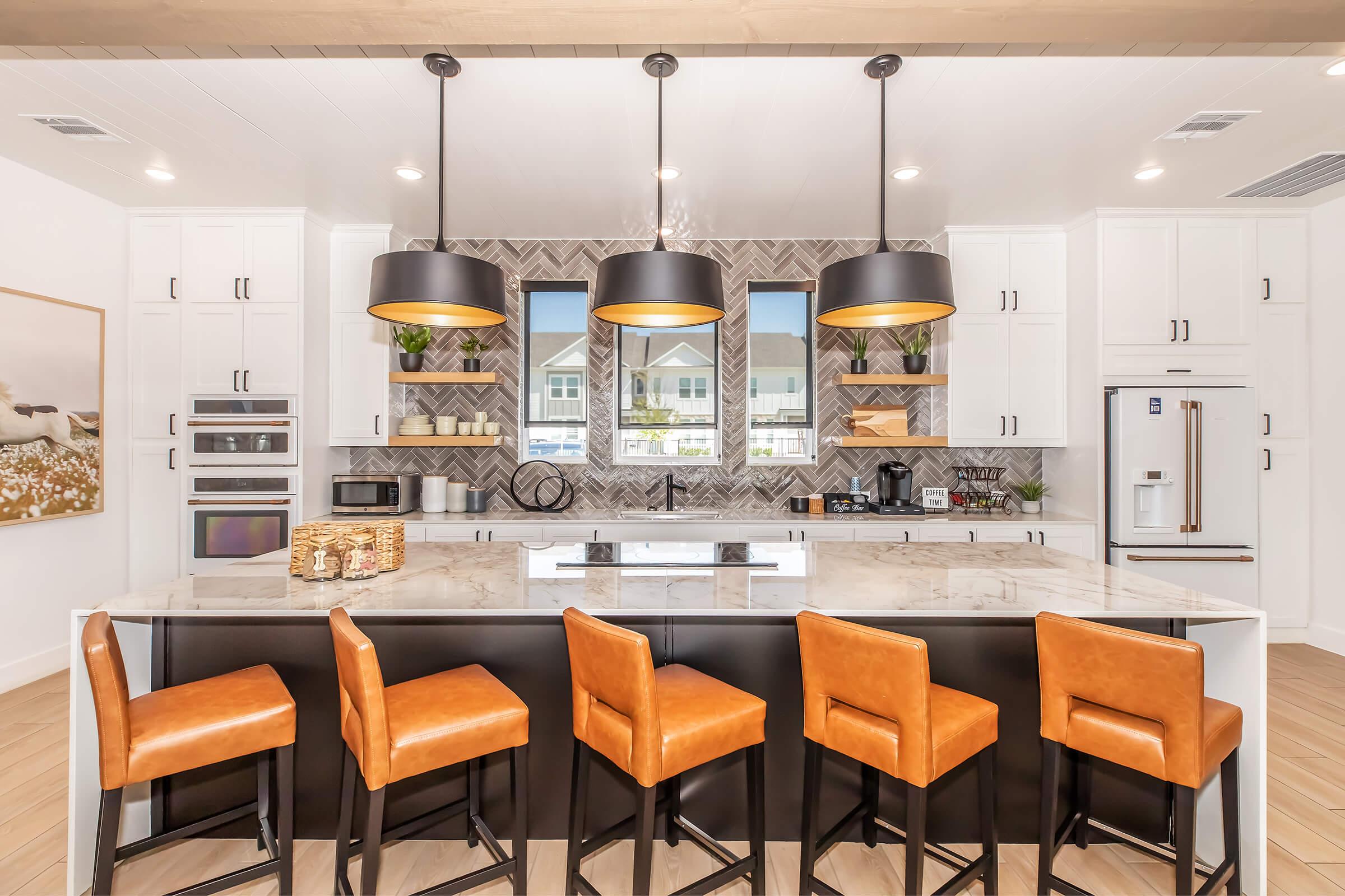 Modern kitchen with a large island featuring a marble countertop. Four brown leather bar stools are arranged around the island. The kitchen has white cabinets, a patterned backsplash, and three large pendant lights hanging above. Large windows illuminate the space, showcasing a bright and airy atmosphere.