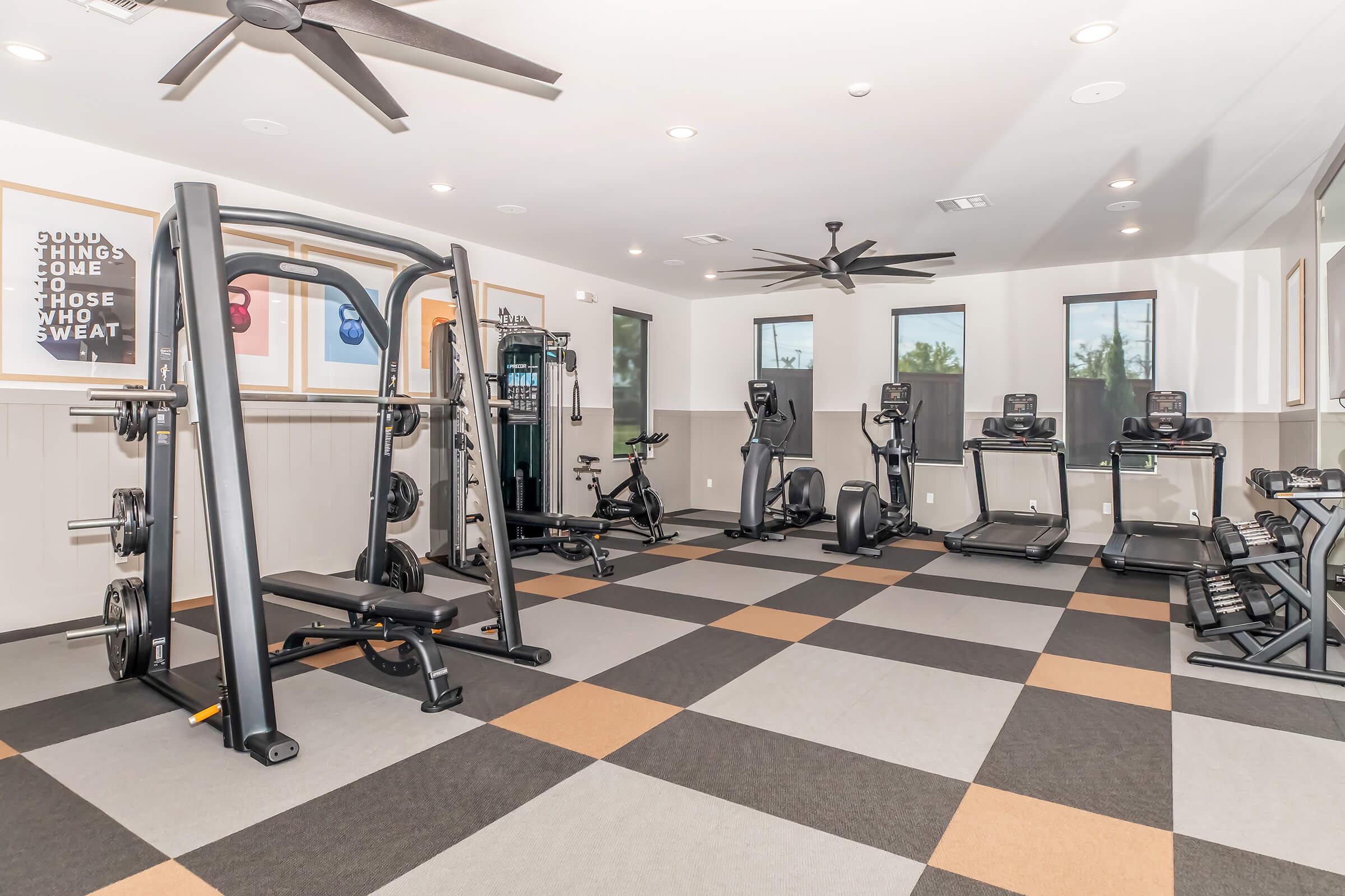 A modern gym interior featuring various exercise equipment, including weight machines, treadmills, and stationary bikes. The floor is covered with a checkered pattern of black, gray, and orange tiles. Large windows provide natural light, and motivational posters adorn the walls.