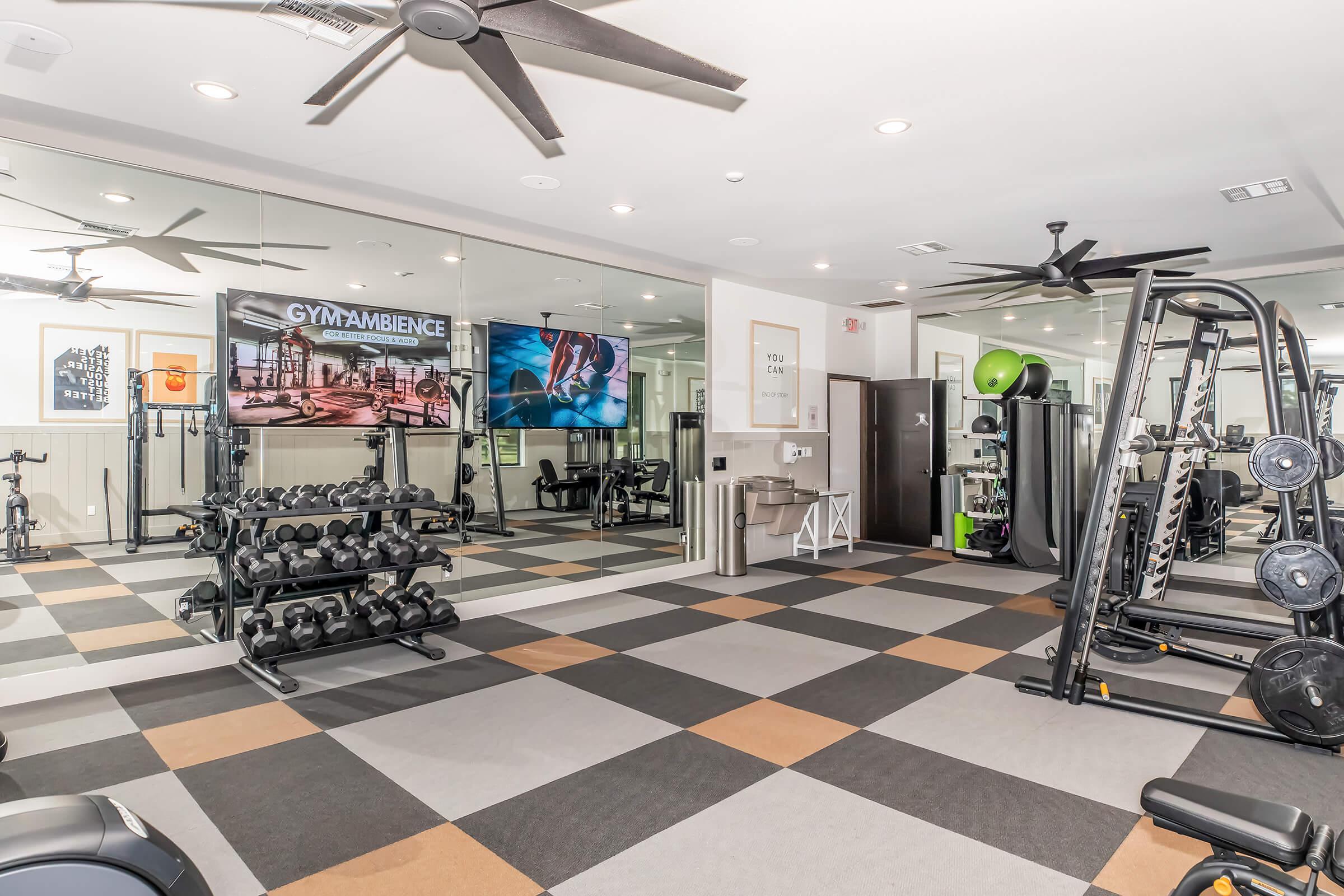 A modern gym interior featuring a mirrored wall, exercise equipment, and weights on racks. There are two televisions displaying fitness-related content, along with large ceiling fans and a variety of workout machines. The flooring consists of cushioned tiles in contrasting colors.