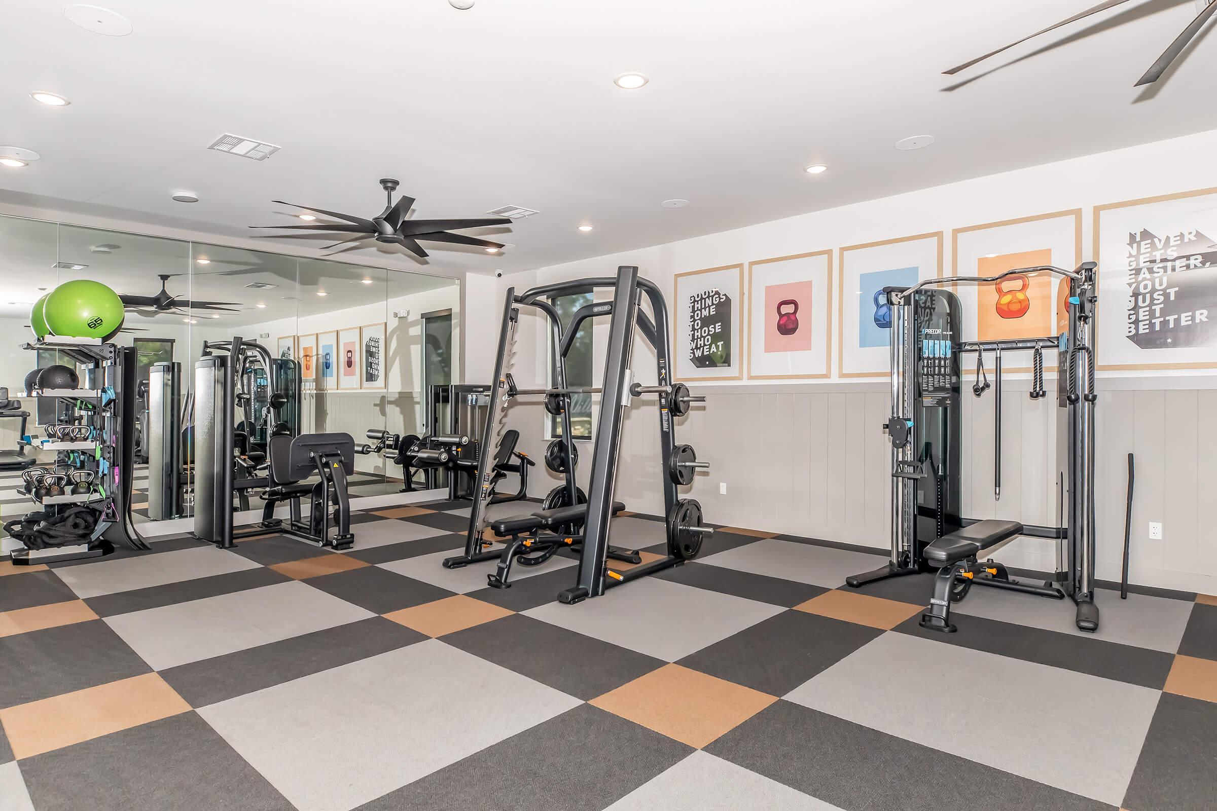 A modern gym interior featuring various fitness equipment, including weight machines and a cable machine. The flooring consists of colorful square tiles, and there are framed motivational posters on the walls. Large mirrors and ceiling fans enhance the spacious feel of the gym.