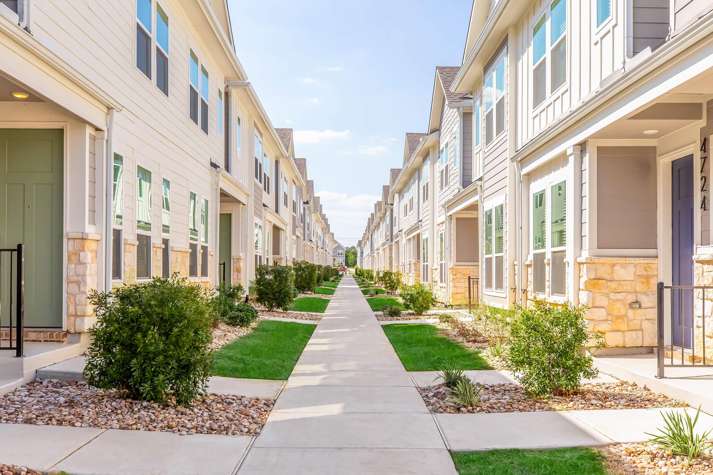 A well-maintained residential area featuring two rows of modern townhouses. Each unit has a porch, and there are neatly trimmed bushes and a landscaped pathway running between the buildings, leading towards the horizon under a clear blue sky.