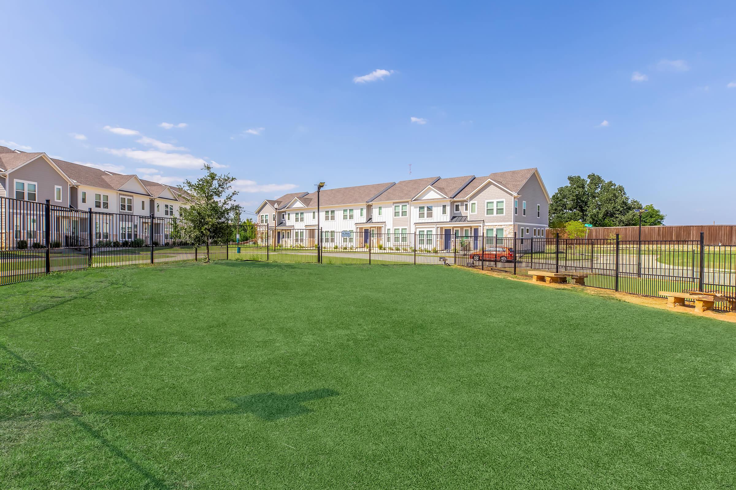 A spacious green lawn enclosed by a black fence, with several modern townhouse buildings in the background. The sky is clear and blue, with a few clouds. There is a red car parked nearby, and the area appears well-maintained, suggesting a community space or recreational area.