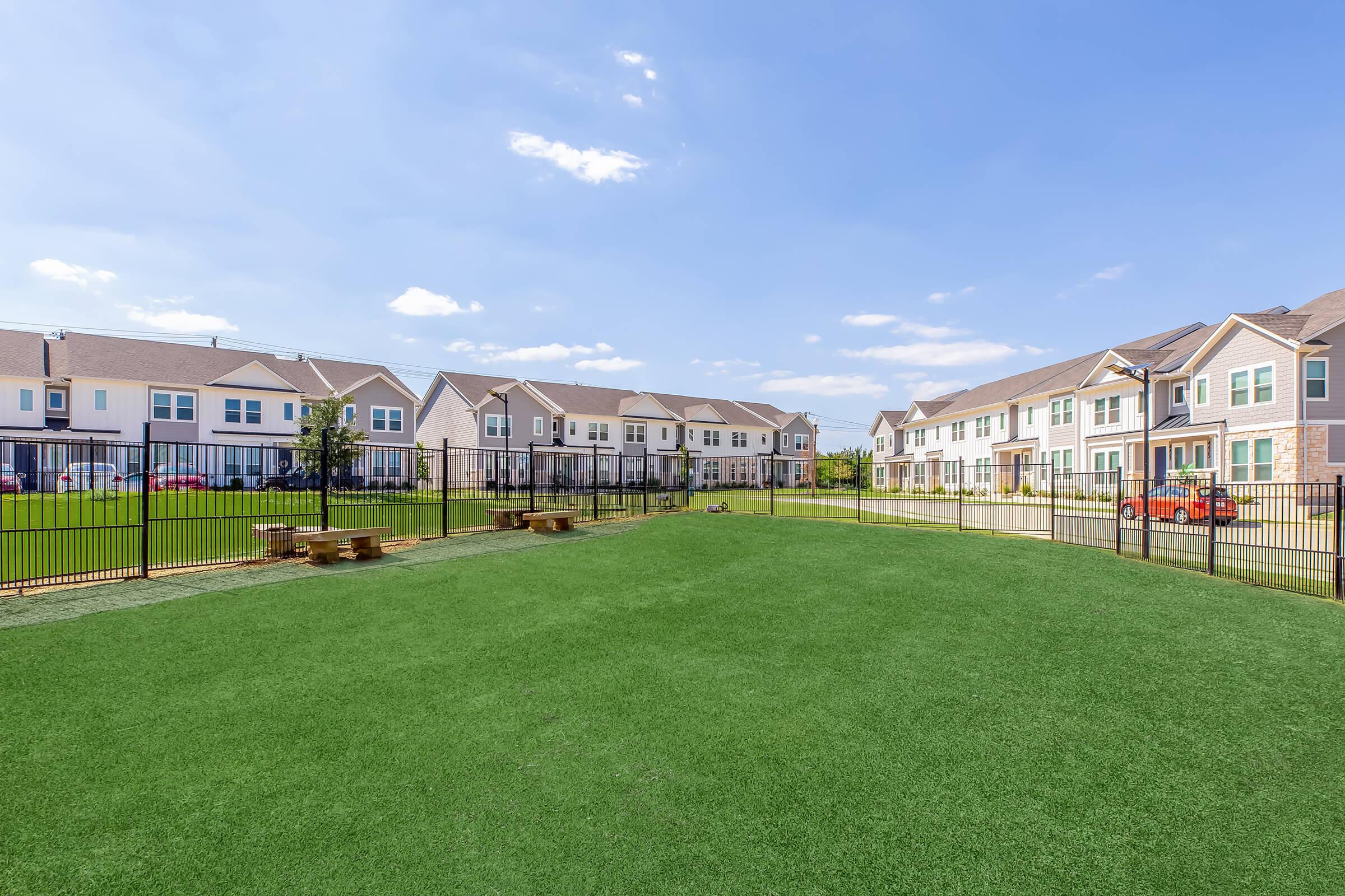 View of a landscaped outdoor area featuring a grassy field surrounded by a black metal fence. In the background, residential buildings with multiple units are visible under a clear blue sky with scattered clouds. The space includes picnic benches, ideal for recreational activities.