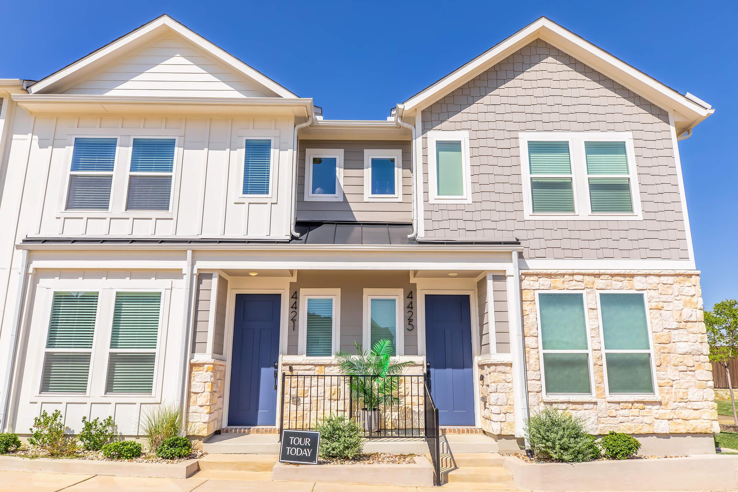 A modern two-story duplex with light-colored siding and a stone facade. The building features four windows on the upper level and two doors on the lower level, each adorned with decorative plants. A sign labeled "TOUR TODAY" is placed in front of the entrance, against a clear blue sky.