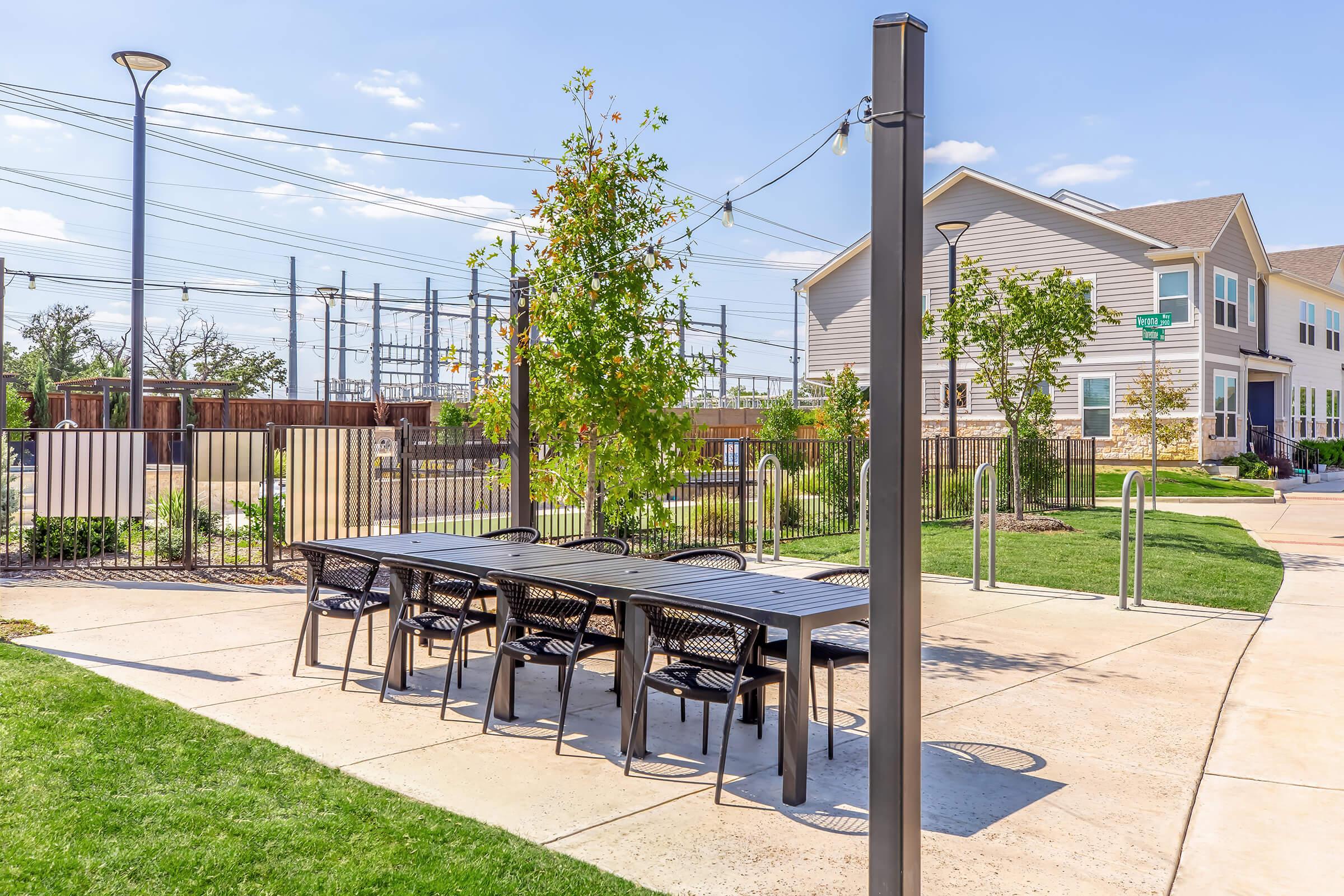 A modern outdoor patio area featuring a long black table with black chairs, surrounded by green grass and trees. In the background, there are power lines and a residential building, along with a fenced area. The scene is set under a clear blue sky, creating a bright and inviting atmosphere.