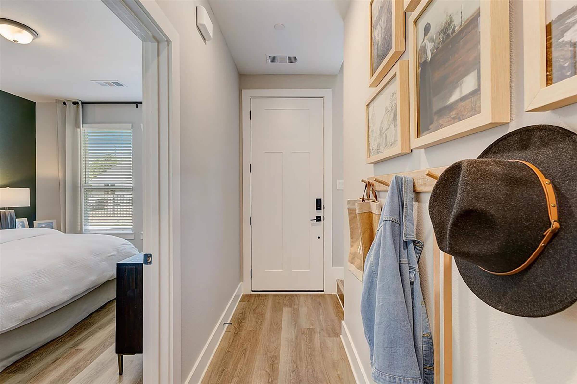 A hallway that leads to a bedroom, featuring light wood flooring and neutral-colored walls. There’s a closed white door, a wall with framed pictures, a coat rack with a hat and a denim jacket hanging, and a glimpse of a neatly made bed in the adjacent room. Natural light filters through a window.