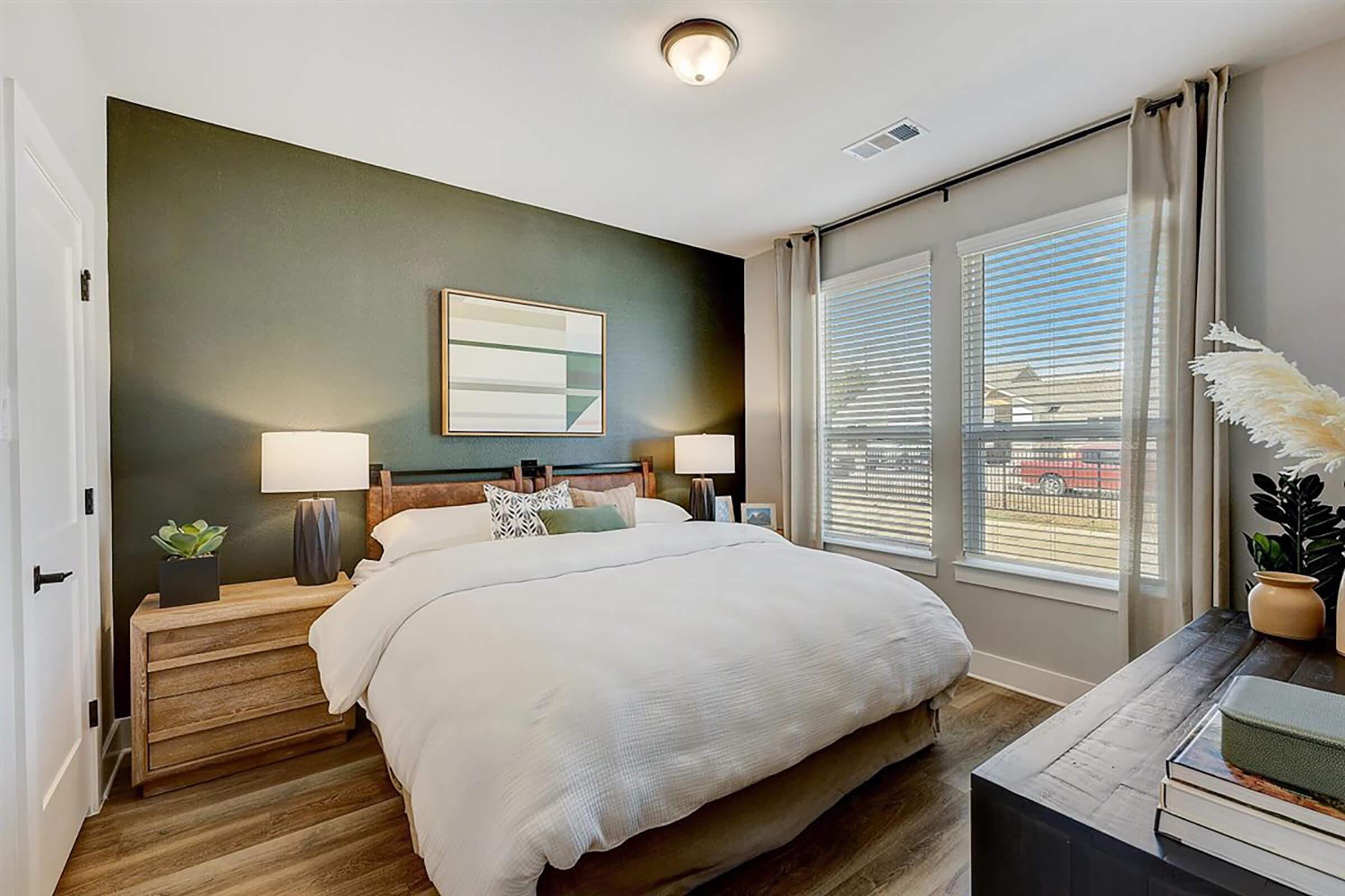 A cozy bedroom featuring a queen-sized bed with a white comforter, two bedside lamps on wooden nightstands, and large windows with sheer curtains. A framed artwork hangs above the bed, and there is a plant on the nightstand, creating a warm and inviting atmosphere.