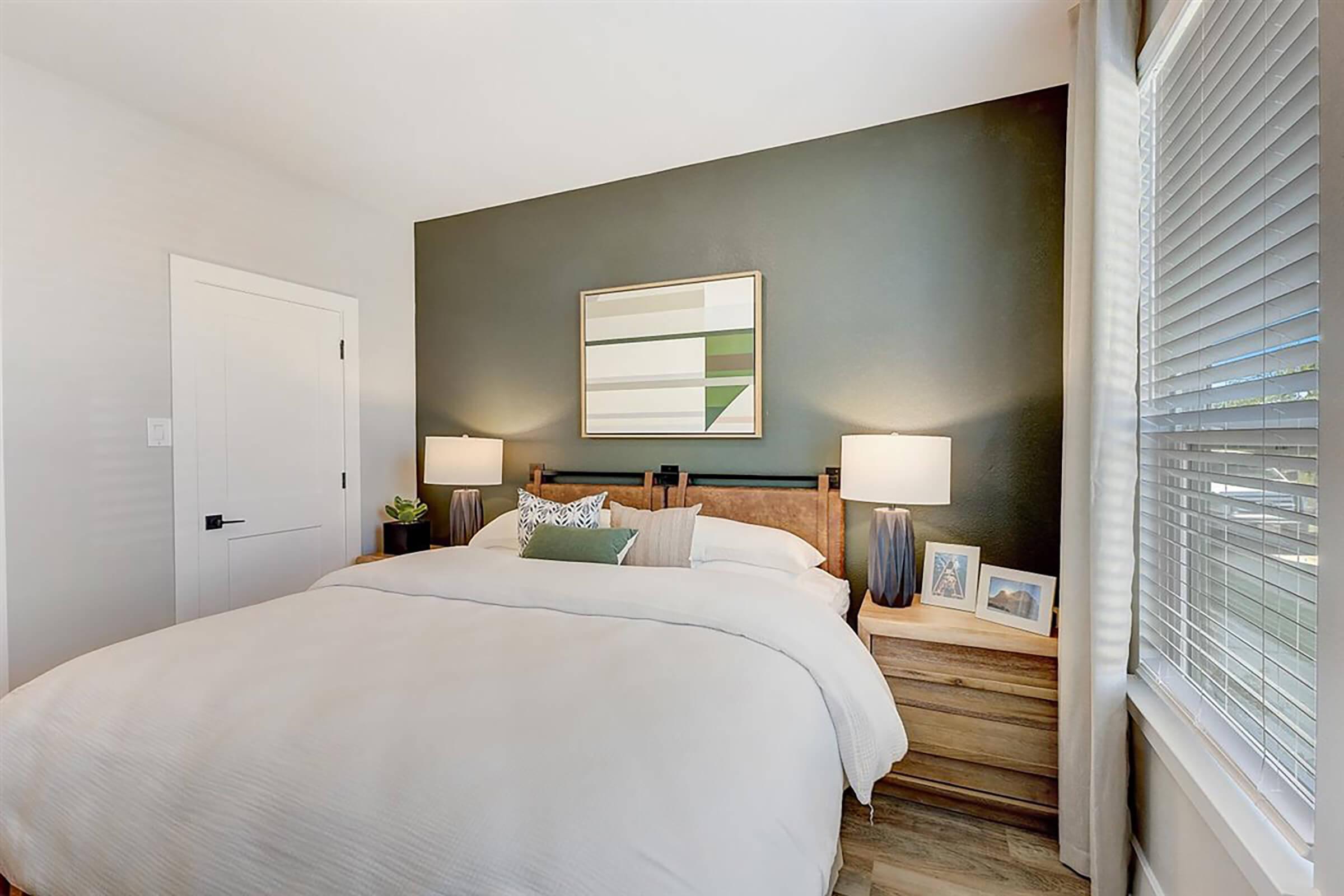 Cozy bedroom featuring a large bed with white bedding, wooden nightstands on either side, and two lamps. A modern art piece hangs above the bed on a dark green accent wall, while natural light filters through window blinds, creating a warm and inviting atmosphere.