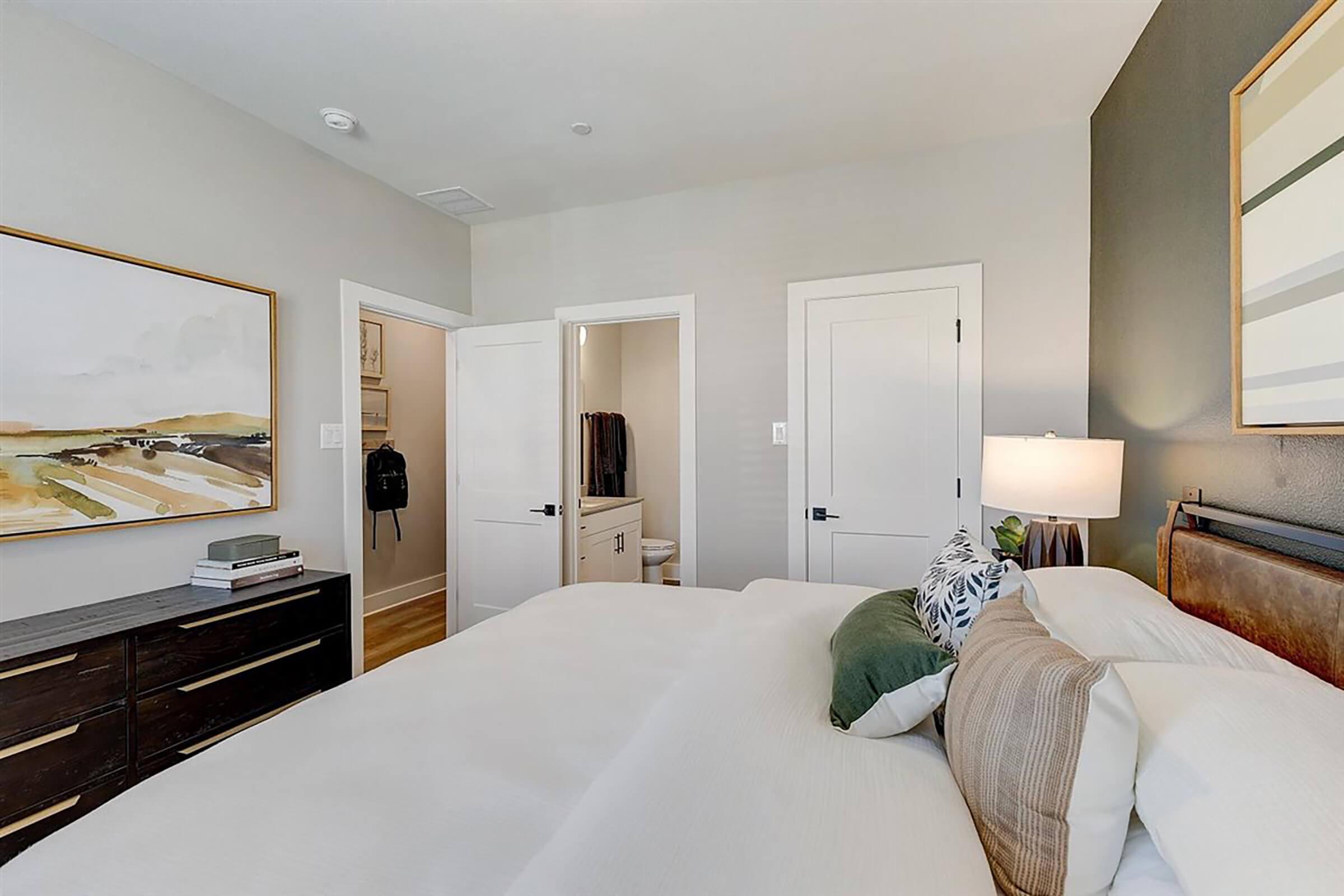 A cozy bedroom featuring a neutral color palette with a large bed adorned with decorative pillows. There's a wooden dresser with books and a lamp beside it. A door leads to a closet, while another door opens to a bathroom. The wall art adds a touch of warmth and character to the space.