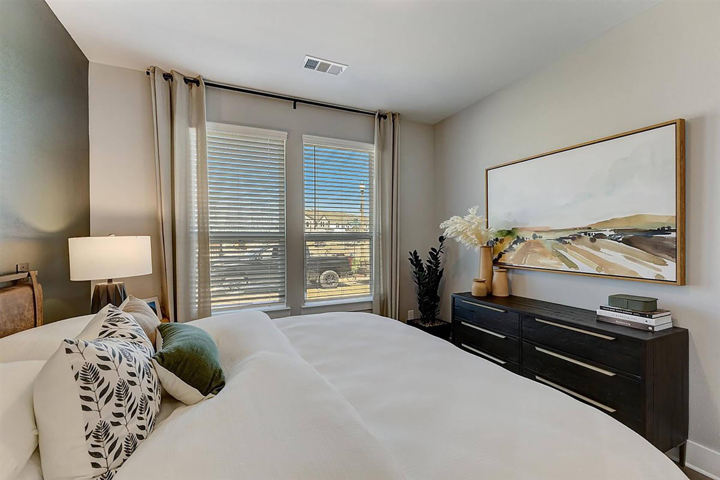 A cozy bedroom featuring a large bed with white bedding and decorative pillows. Natural light streams through two windows adorned with light curtains. A stylish side table with a lamp and a dark wooden dresser enhance the room's decor, while a piece of landscape artwork hangs on the wall.