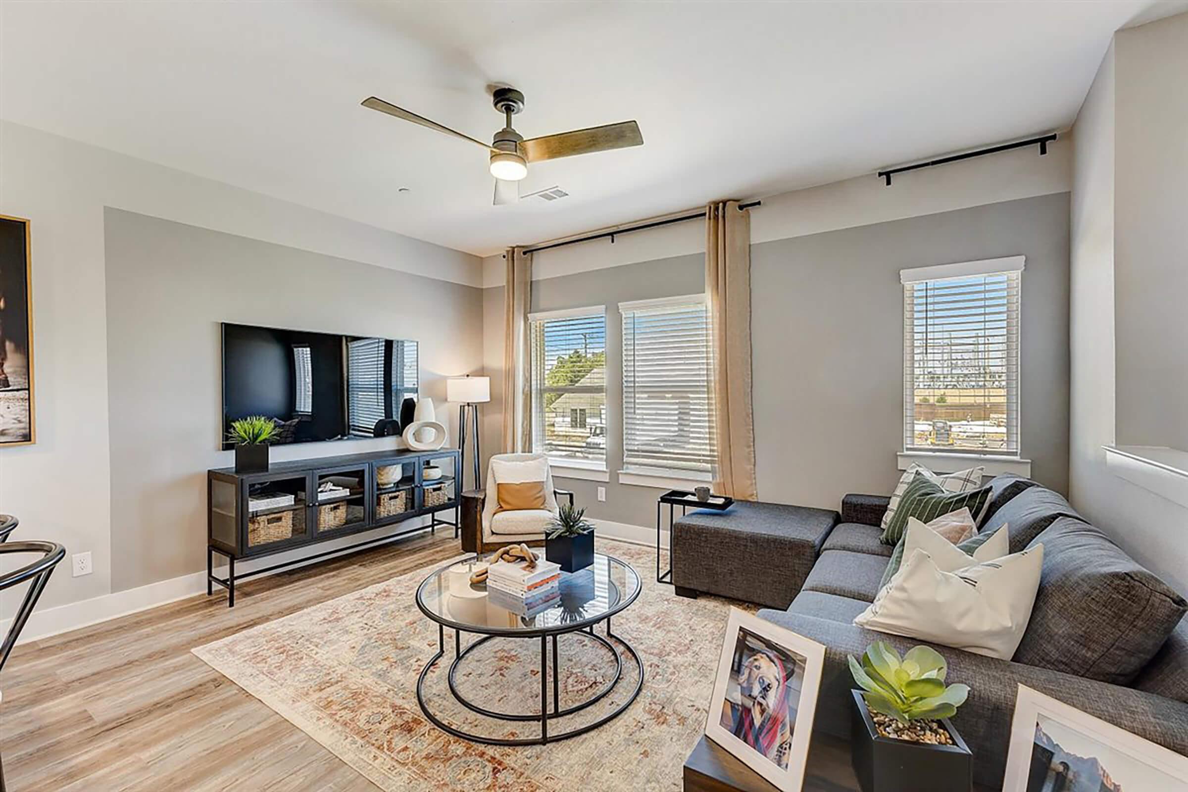 A modern living room featuring a gray sectional sofa, a round coffee table with decorative items, a wall-mounted TV, and large windows with natural light. The space includes a potted plant, decorative art on the wall, and a cozy area rug, creating a warm and inviting atmosphere.