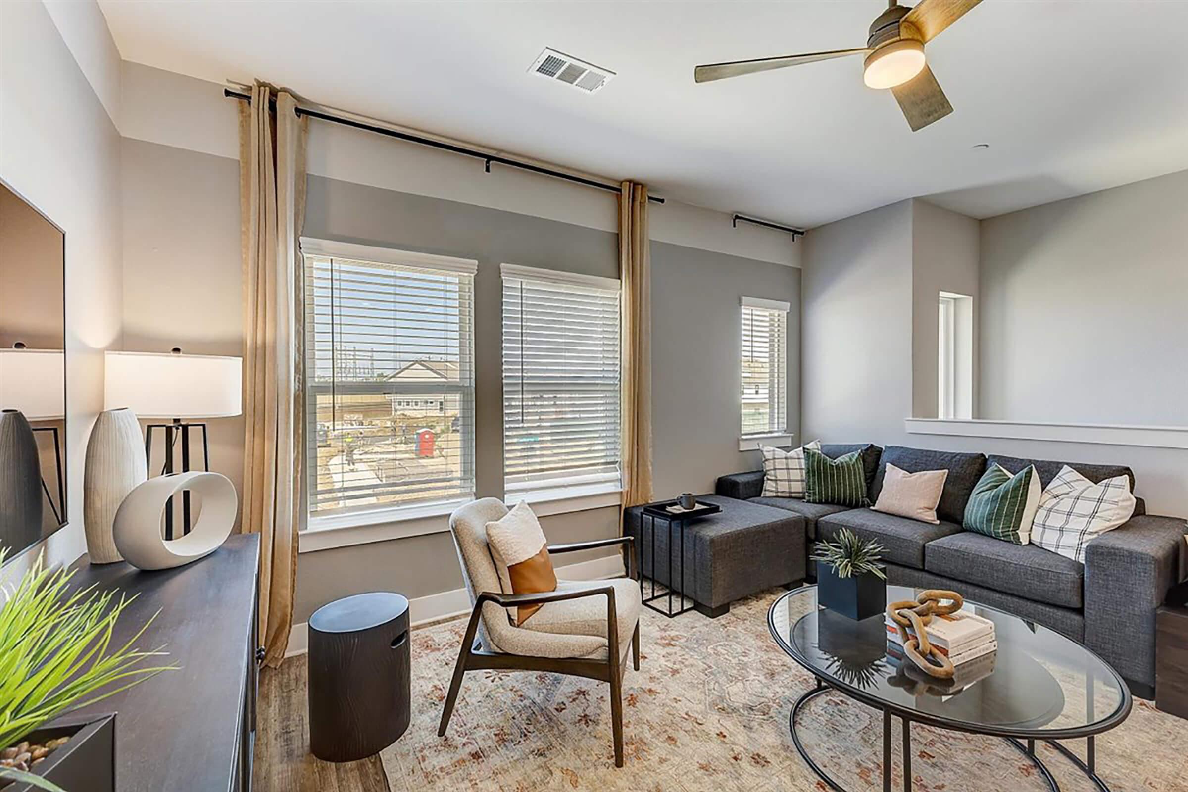 Modern living room featuring a gray sectional sofa with decorative pillows, a round glass coffee table, and a stylish armchair. Natural light streams through large windows with blinds. The decor includes a geometric area rug and minimalistic accessories, creating a cozy and inviting atmosphere.