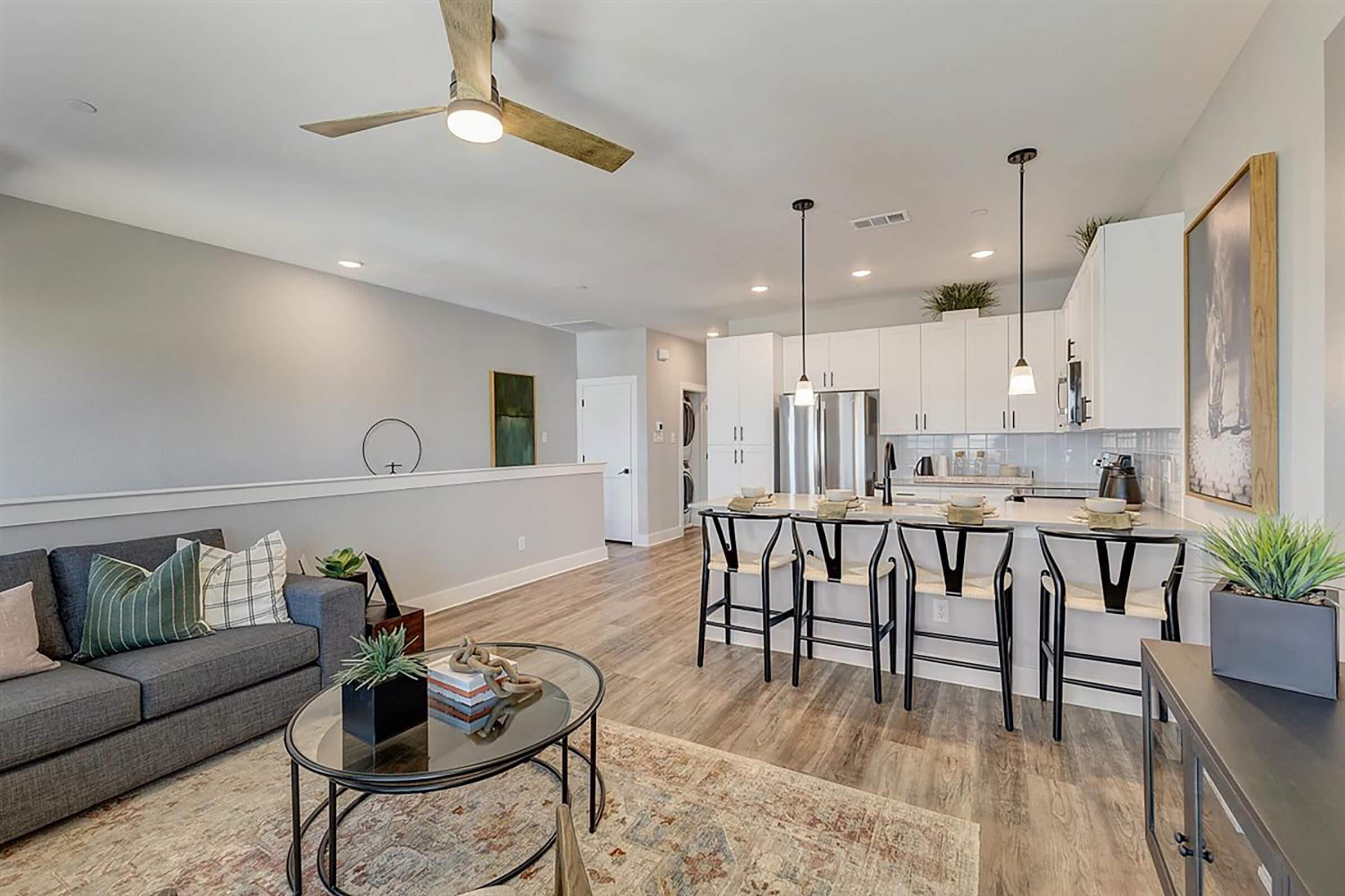 Modern open-concept kitchen and living room featuring a gray sofa with decorative pillows, a round glass coffee table, a light-colored dining area with four black chairs, and contemporary cabinetry. Large windows allow natural light, and subdued decor adds a cozy ambiance.