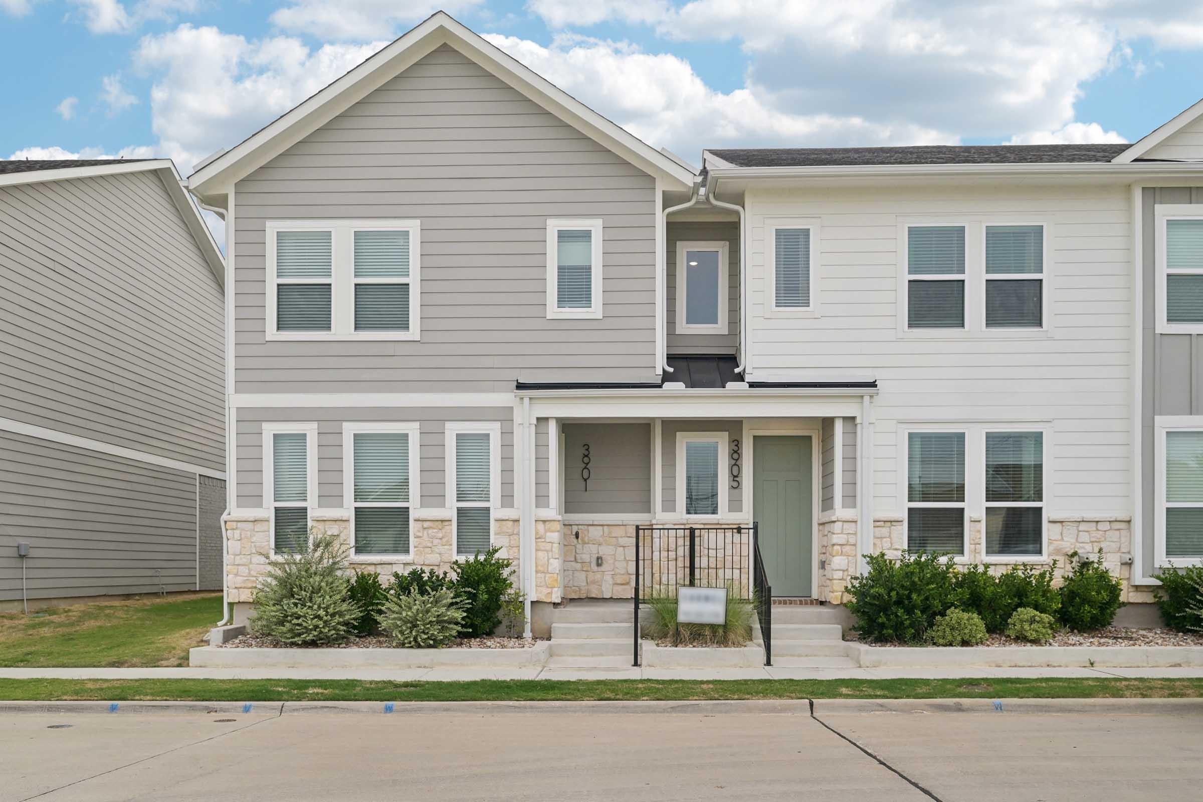 A modern residential building featuring two attached homes with a light gray and white exterior. Each unit has multiple windows, a porch with steps, and landscaped front yards with shrubs. The setting includes a clear blue sky with fluffy clouds.