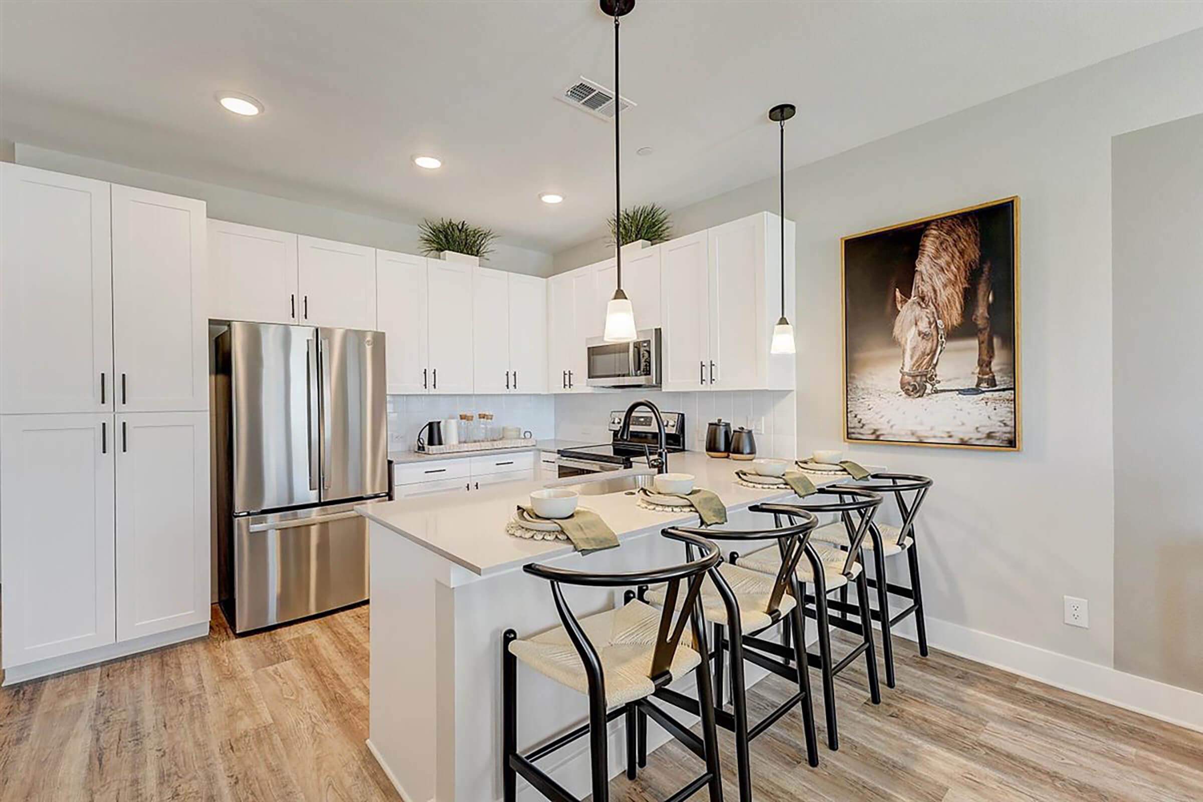 Modern kitchen featuring white cabinetry, stainless steel appliances, and an island with seating for four. A large artwork of a horse hangs on the wall, adding a rustic touch. The space is well-lit with recessed lighting and has a warm, inviting atmosphere. Wood-style flooring complements the overall design.