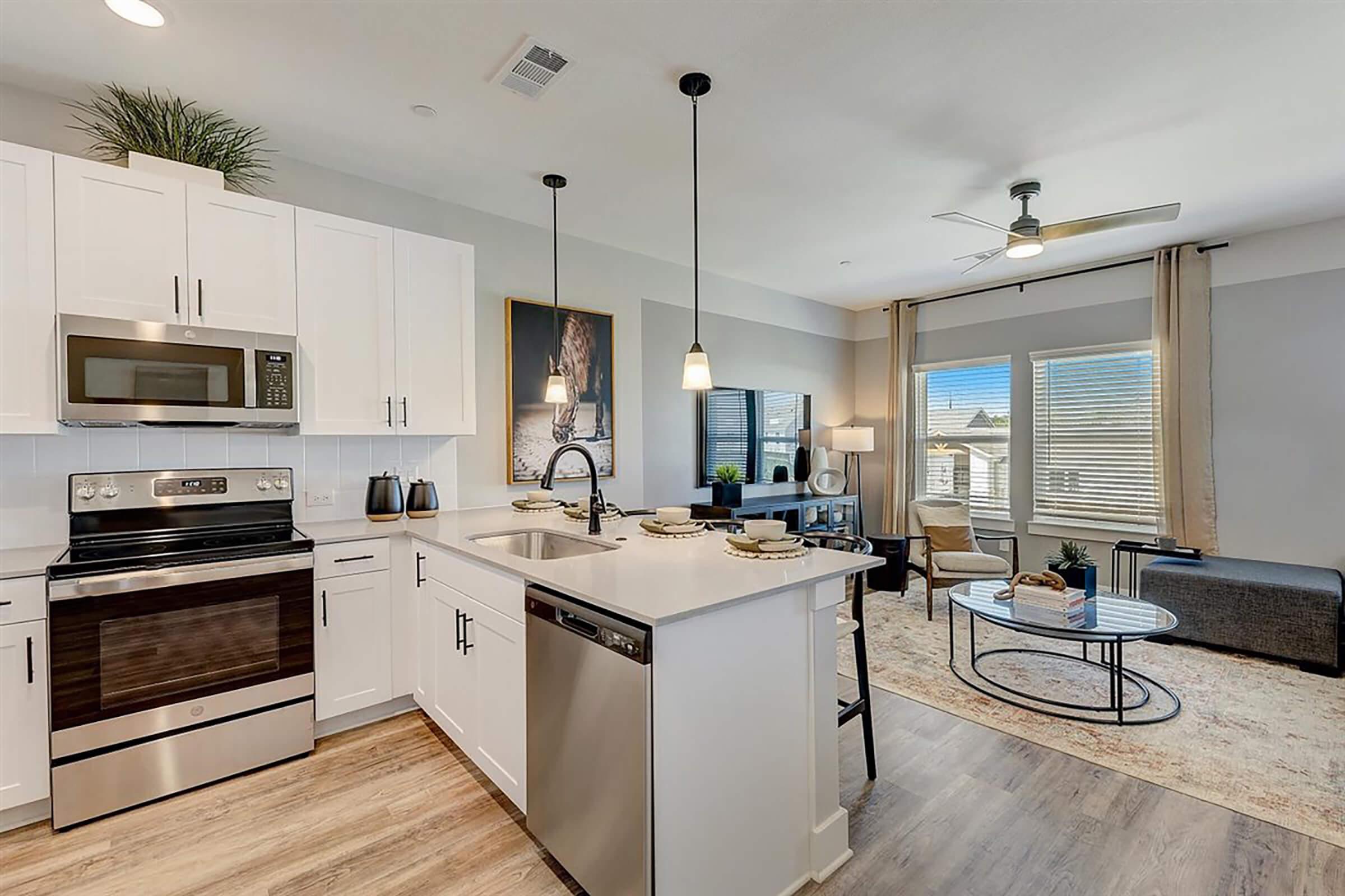 Modern kitchen and living area featuring stainless steel appliances, a center island with seating, and a cozy lounge space with a light-colored rug and a sofa. Large windows allow natural light, while tasteful decor adds a contemporary touch.