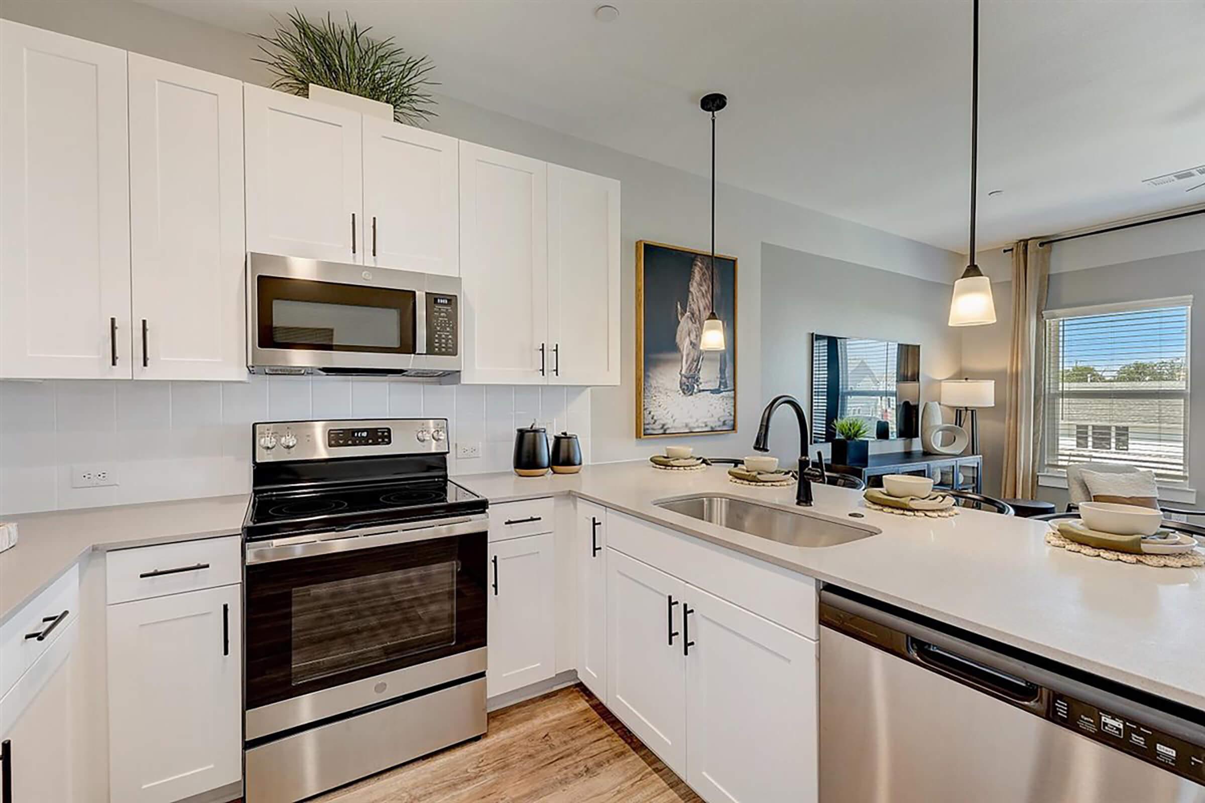 Modern kitchen featuring white cabinets, stainless steel appliances including an oven and microwave, and a sink with a black faucet. The countertop is light-colored, and decorative plates are arranged on the counter. The background shows a cozy living area with large windows and stylish decor.
