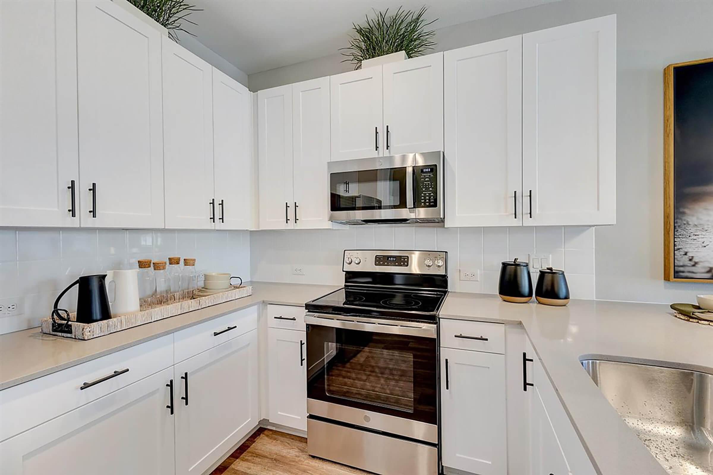 Modern kitchen with white cabinetry, stainless steel microwave, and stove. A wooden countertop displays coffee containers and a bowl, alongside a black kettle. The sink area is visible, with decorative plants on top cabinets. Natural light brightens the space, creating a clean and inviting atmosphere.