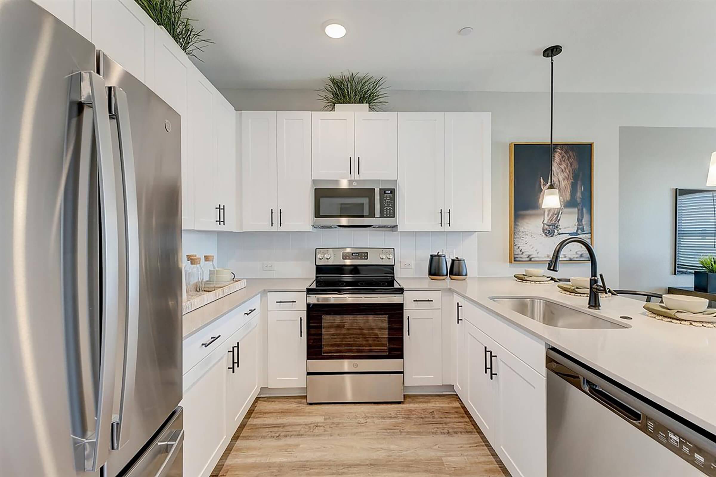 Modern kitchen featuring stainless steel appliances, including a refrigerator, microwave, and oven. The cabinetry is white with a light countertop. A double sink is visible, and decorative elements like plants and dishware enhance the aesthetics. Natural light floods the space, creating a welcoming atmosphere.