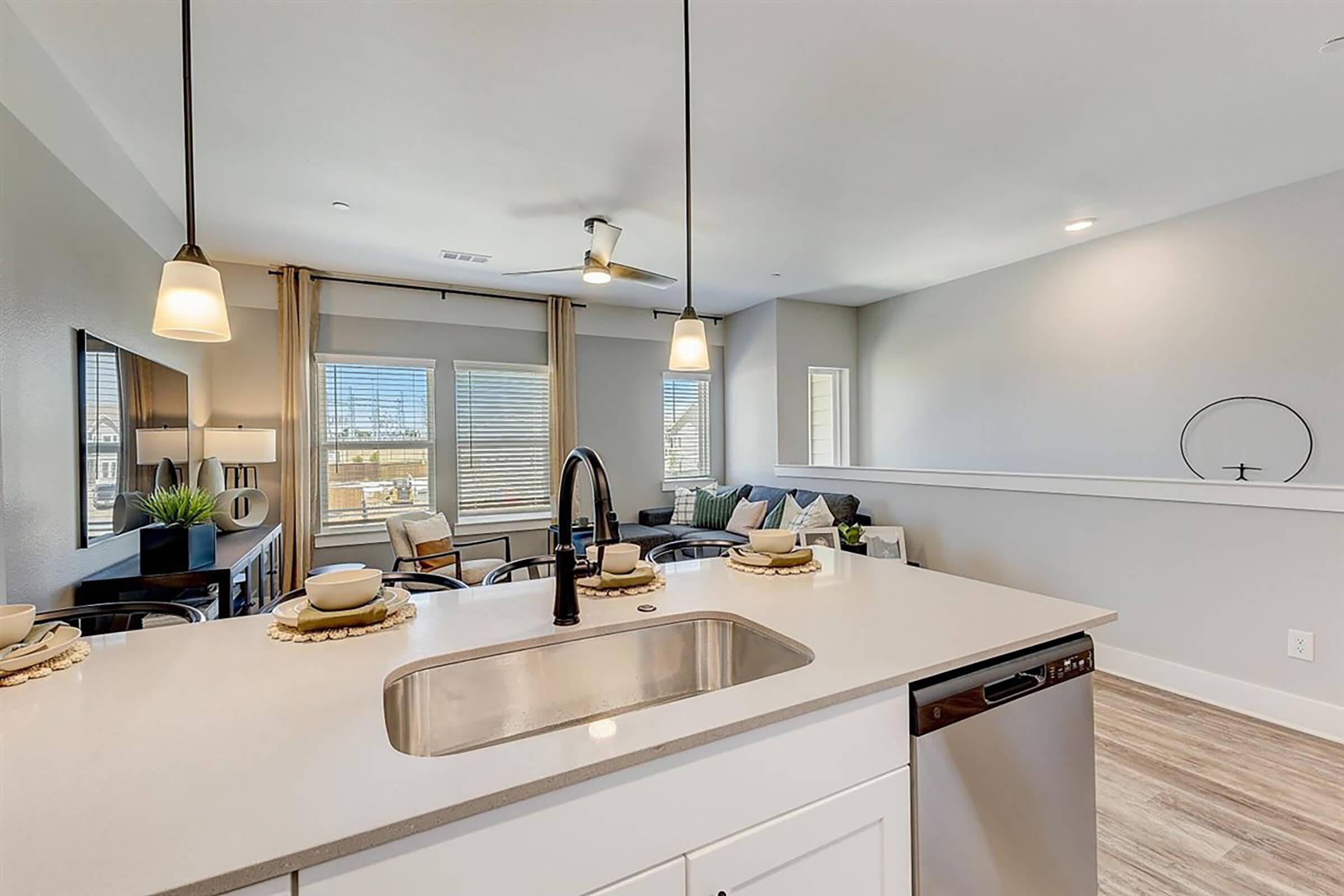 A modern kitchen features a sleek countertop with a sink and a dishwasher. In the background, a cozy living area is visible with large windows letting in natural light, showcasing comfortable seating and decorative touches. Pendant lights hang above the kitchen island, enhancing the contemporary design.