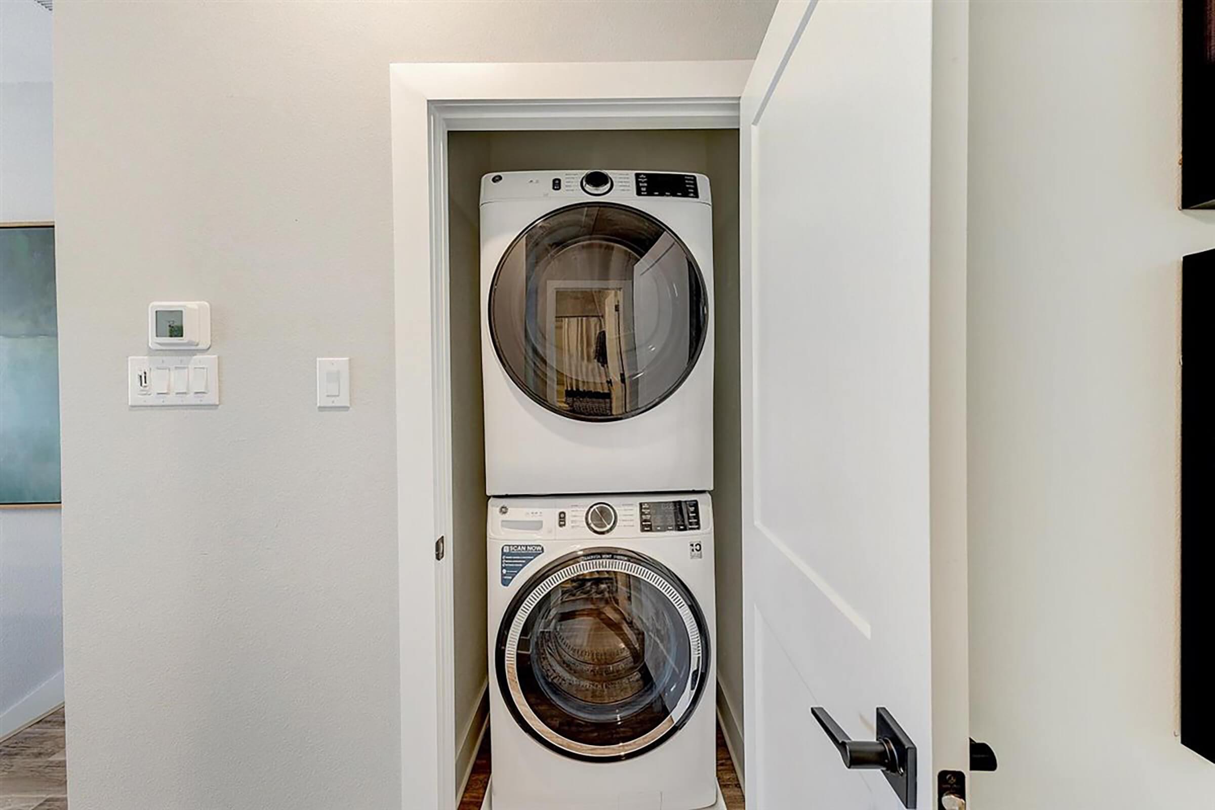 A stackable washer and dryer unit located in a narrow laundry closet with a white door. The walls are painted a light color, and there is a light switch and thermostat visible on the wall beside the unit. The space appears clean and modern.