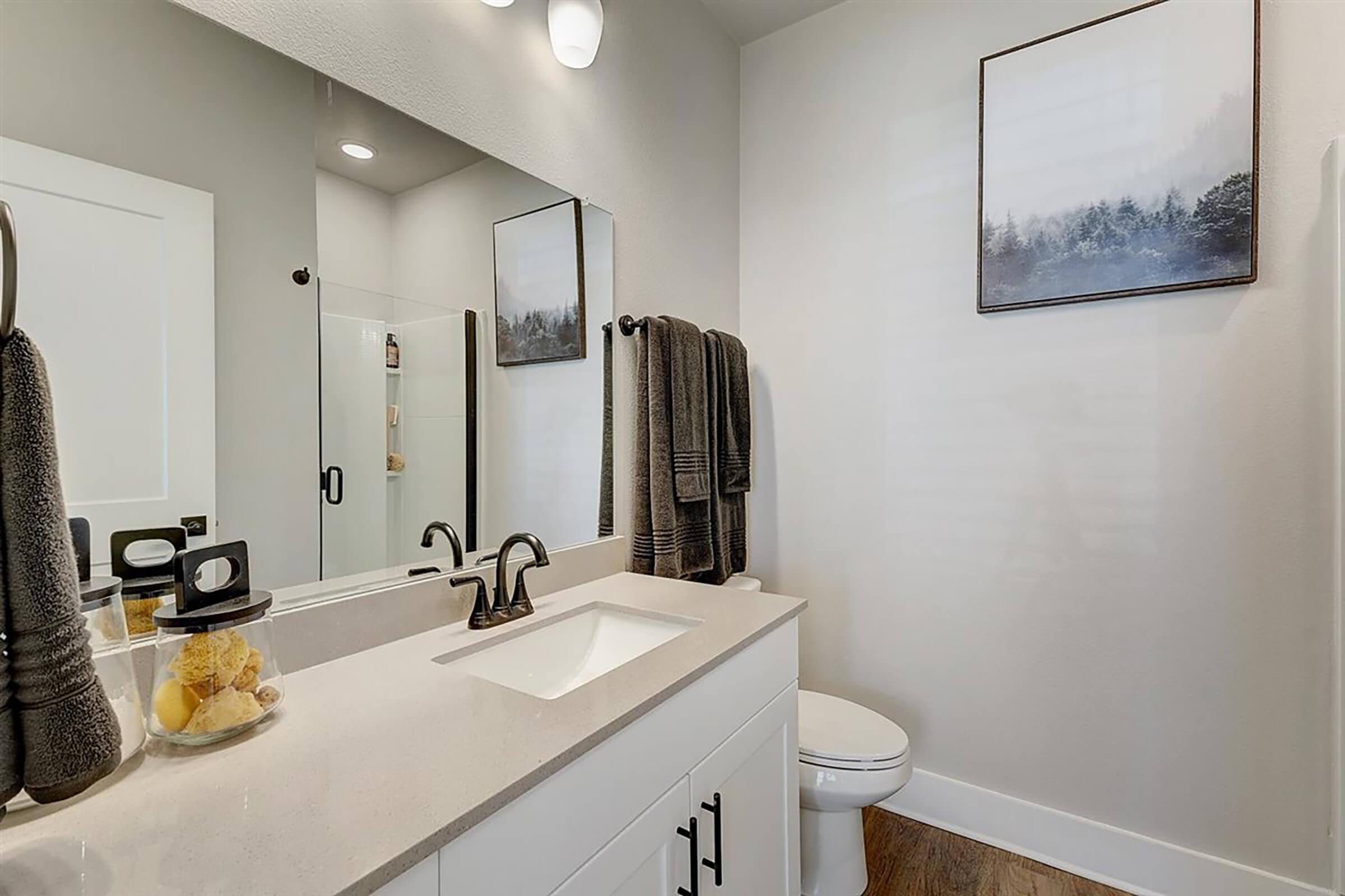 A modern bathroom featuring a white vanity with a sink, dark gray towels hanging on a rack, a framed landscape picture on the wall, and a separate shower. Natural light illuminates the space, highlighting the clean and contemporary design.