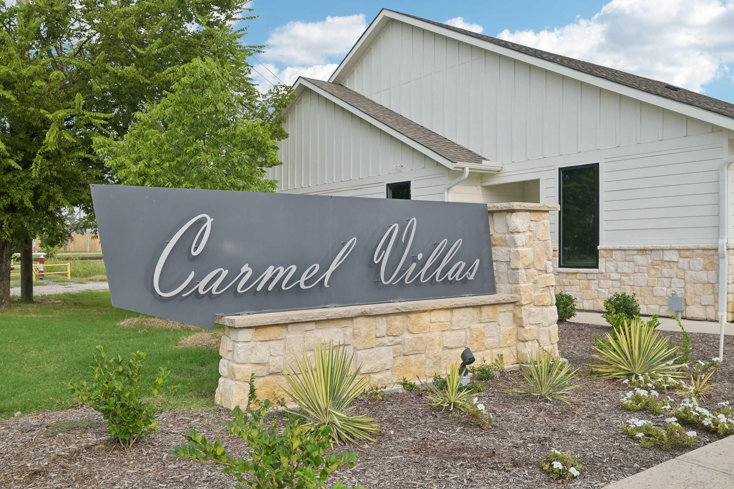 Sign for Carmel Villas in front of a modern building, featuring stylish curved lettering on a dark panel. The surrounding area includes green grass, shrubs, and small plants, with a clear blue sky and scattered clouds in the background.