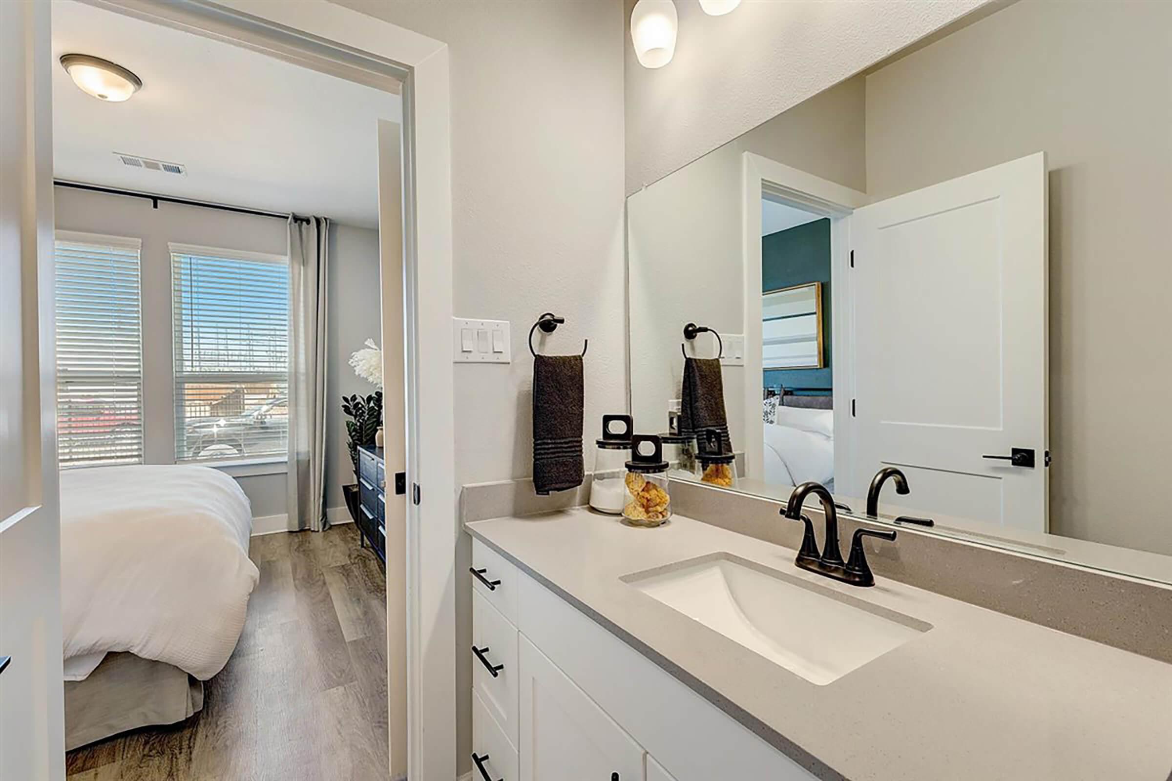 A well-lit modern bathroom featuring a double-sink vanity with a quartz countertop, dark faucet fixtures, and towels hanging on nearby racks. A large mirror reflects the space. The background shows a cozy bedroom with a bed and window, allowing natural light to enter.