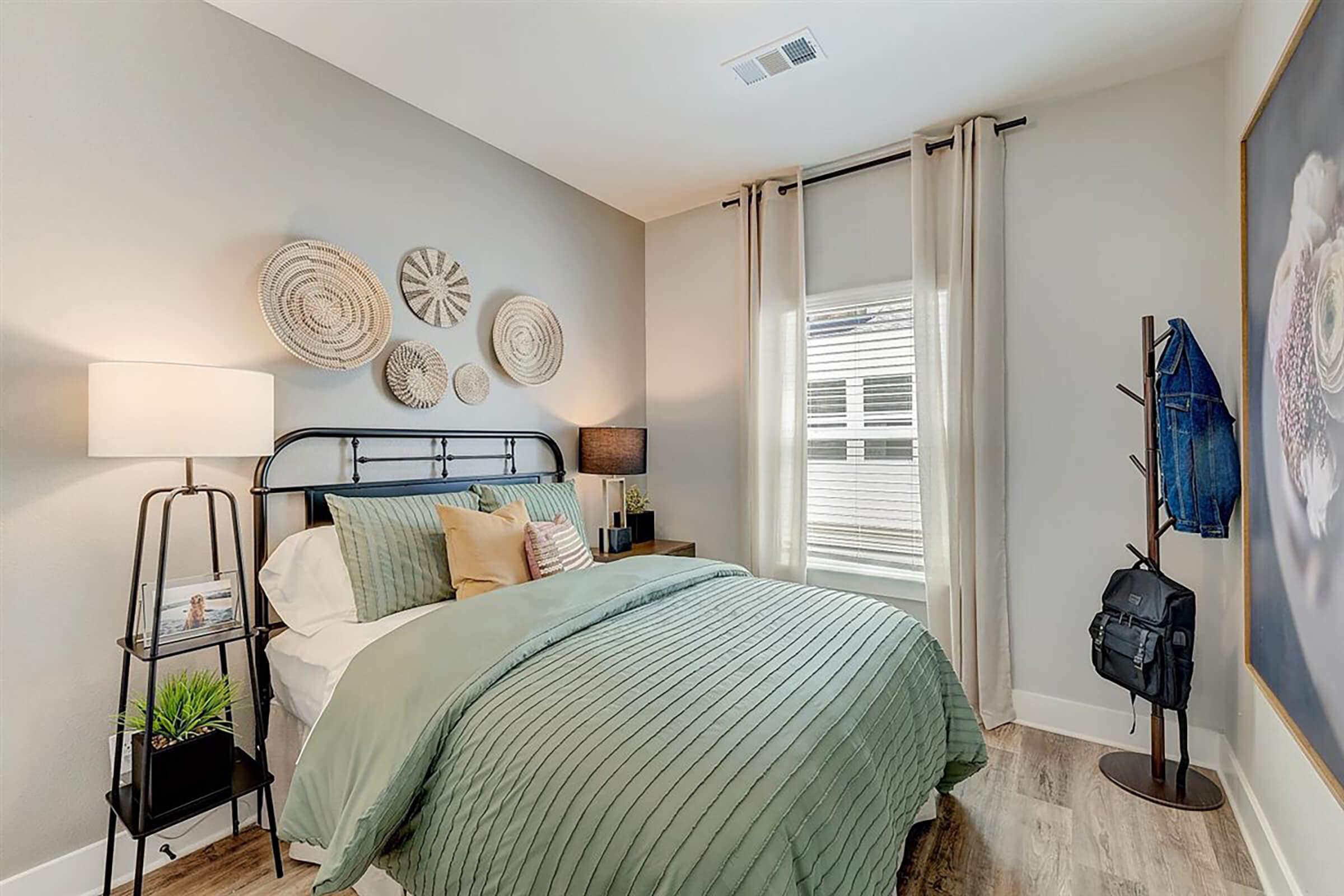 A cozy bedroom featuring a black metal bed frame with a green comforter and decorative pillows. The room includes a bedside lamp on a black stand, wall decor made of woven materials, a window with curtains, and a small coat rack with a backpack. The flooring is wood, adding warmth to the space.