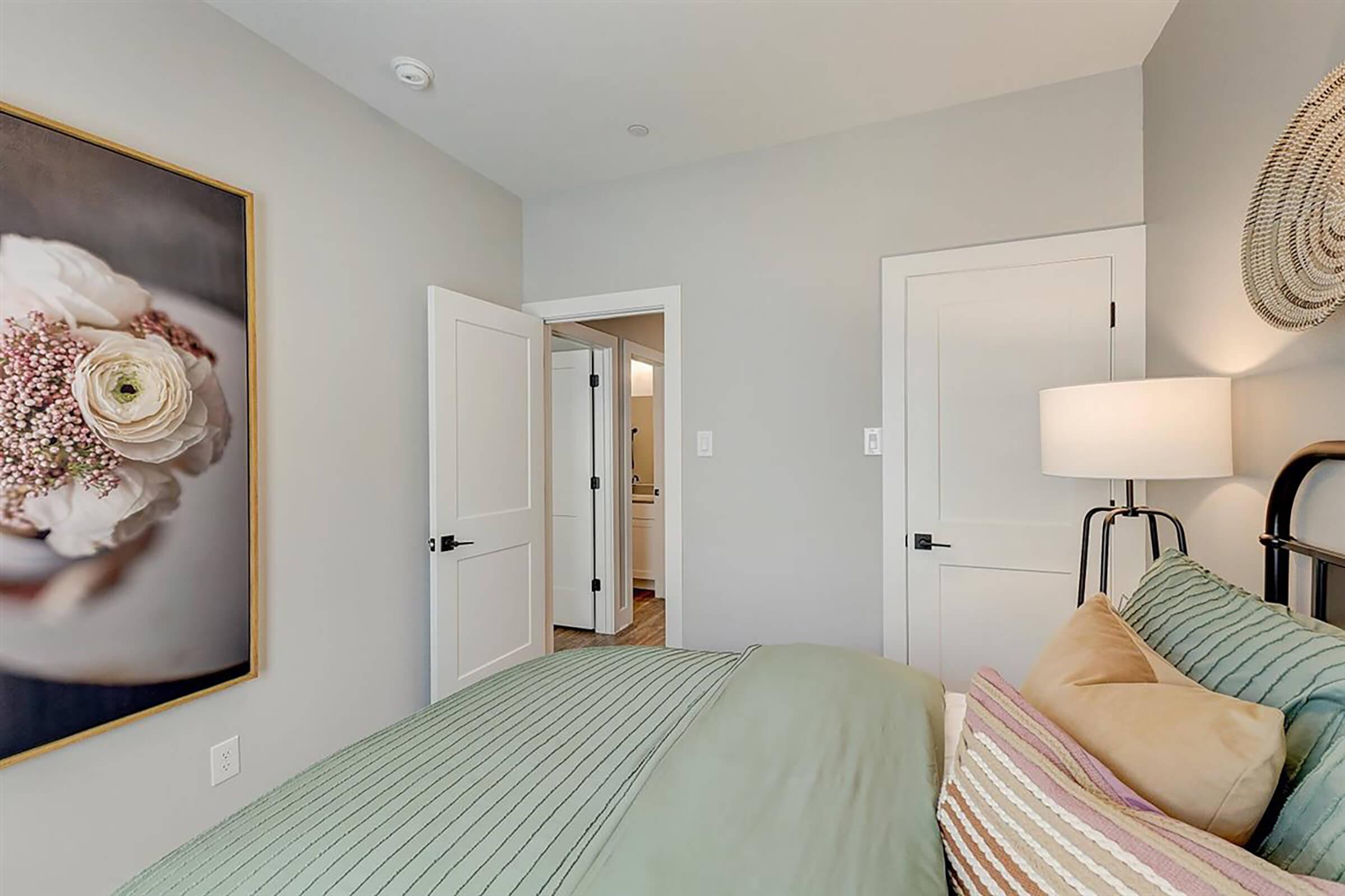 A cozy bedroom featuring a neatly made bed with green and pink bedding. A decorative wall art piece with flowers hangs above the bed. There are two white doors leading to another room and a bathroom. A lamp stands beside the bed, illuminating the space with a warm glow, and the walls are painted in soft gray.