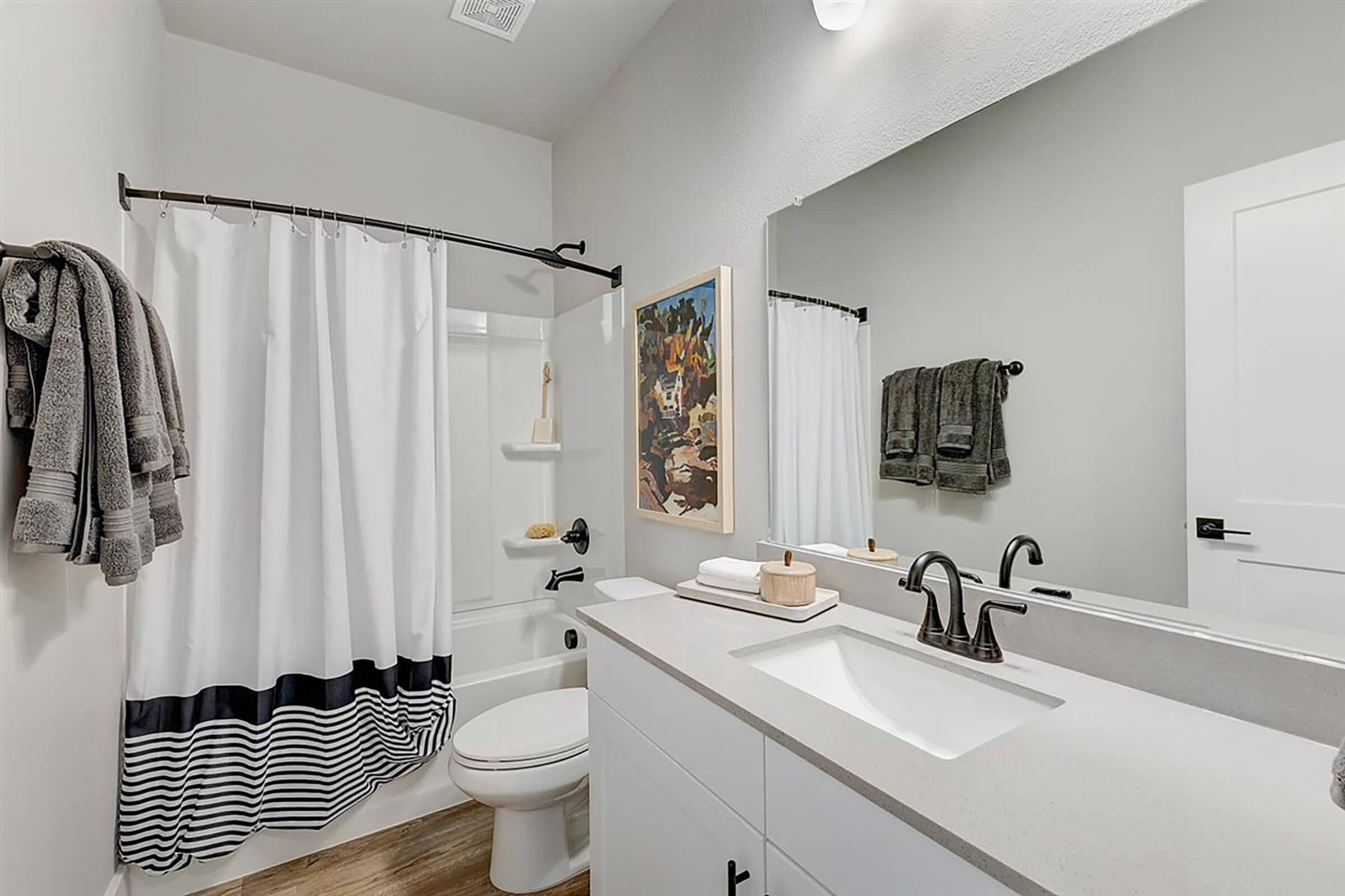 A modern bathroom featuring a white bathtub with a shower curtain, a sleek double sink vanity with black fixtures, a large mirror, and decorative towels. The walls are painted in neutral tones, and there is a colorful piece of art on the wall, adding a touch of personality to the space.