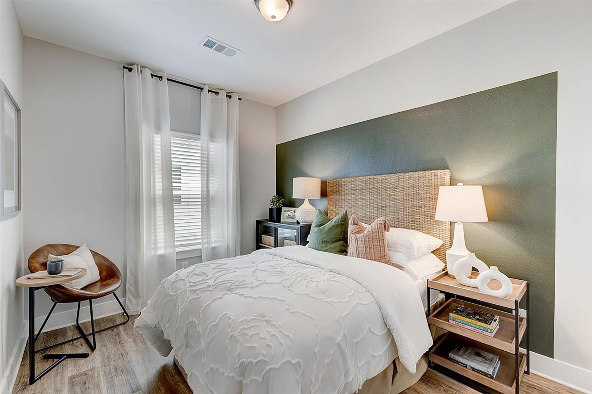 Cozy bedroom featuring a double bed with a white quilted cover, decorated with green and beige accent pillows. A bedside table with a lamp is on each side, and natural light filters through sheer curtains. A stylish armchair and decorative items complement the modern decor.