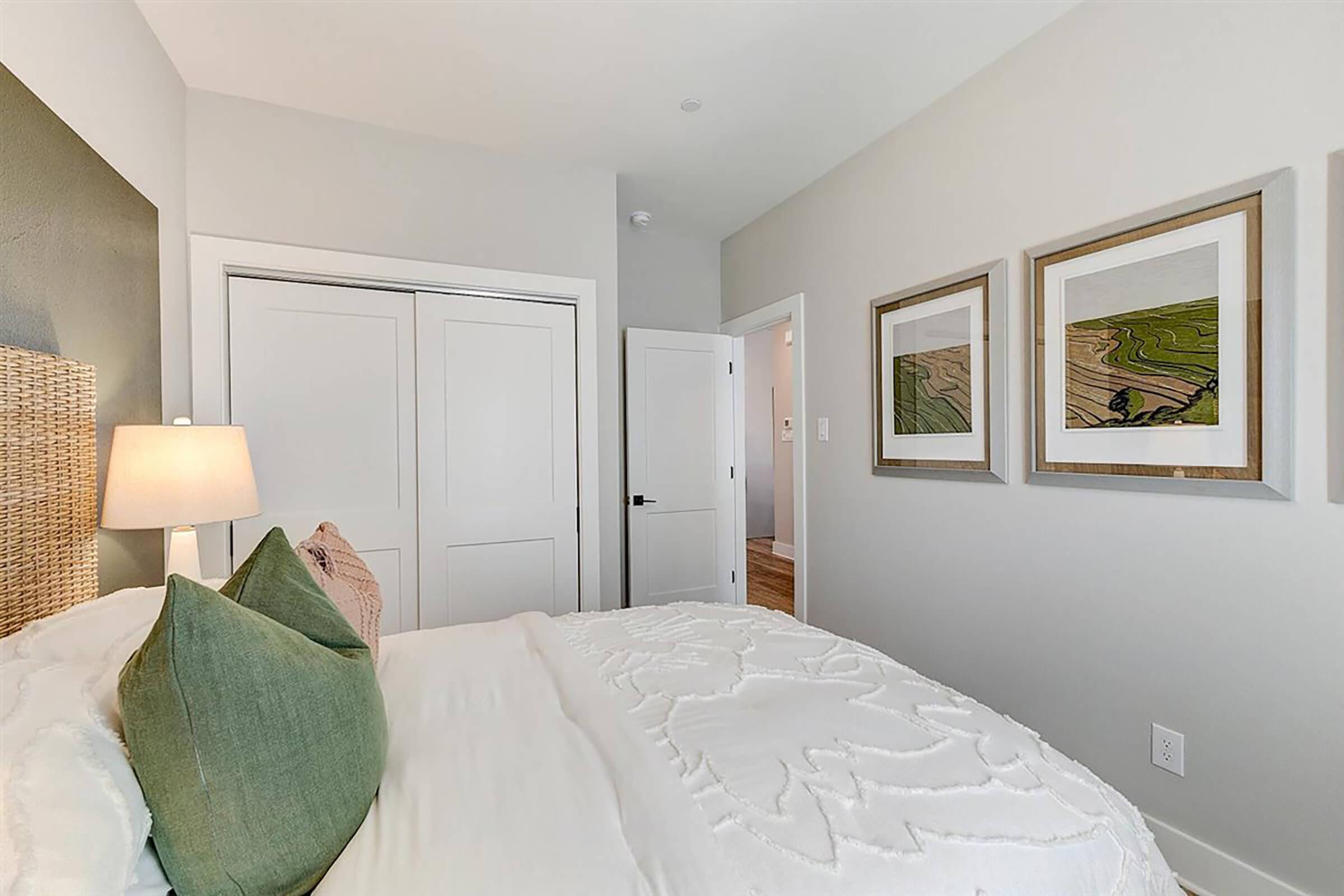 Cozy bedroom featuring a neatly made bed with decorative pillows, a bedside lamp, and a textured headboard. Two framed artworks hang on the wall, and a closet door is visible. The room has a light color scheme with modern decor and a welcoming atmosphere.