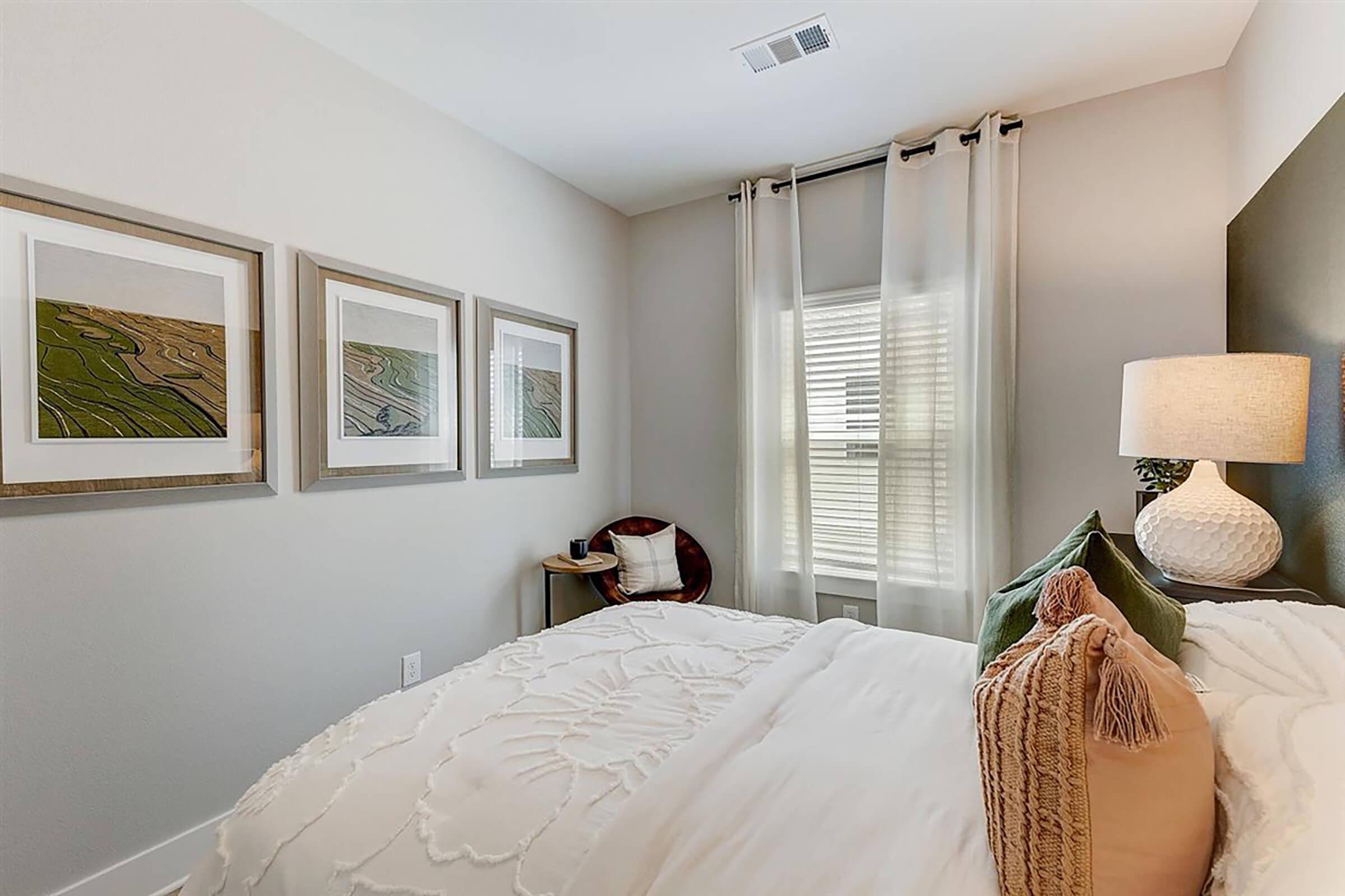 A cozy bedroom featuring a neatly made bed with decorative pillows, framed artwork on the wall depicting landscapes, a lamp on a bedside table, and light curtains allowing natural light to filter in through the window. The walls are painted in a soft, neutral tone, creating a calm atmosphere.