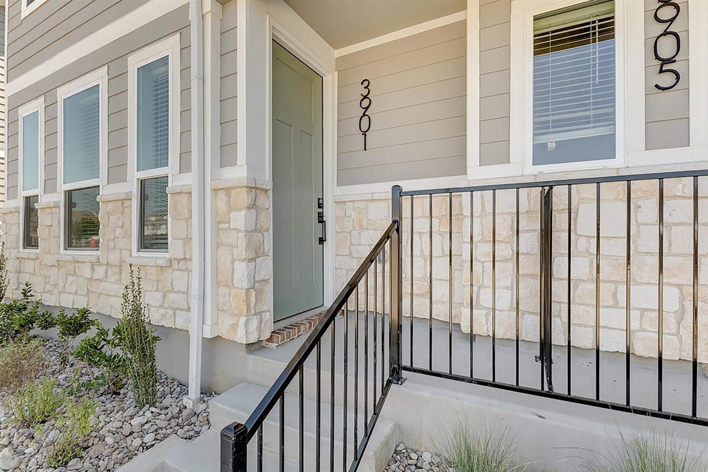 Entrance to a modern home featuring a light green door, stone accents on the lower wall, and a black metal railing. The home has large windows on either side of the door. The pathway is lined with small rocks and plants, creating an inviting entryway.