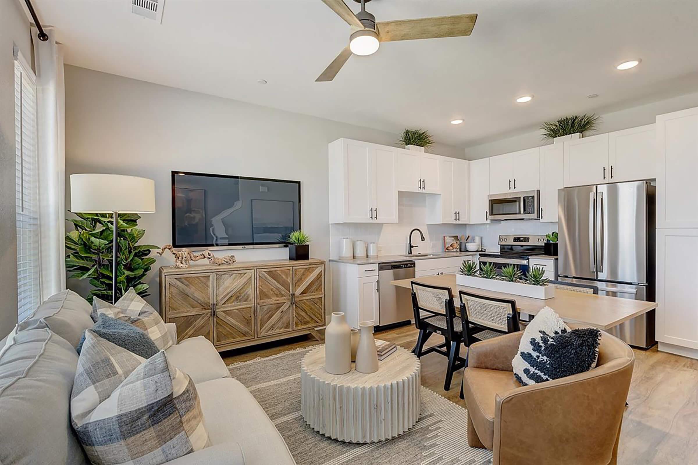 A modern kitchen and living area featuring white cabinetry, stainless steel appliances, a wooden dining table with chairs, a decorative vase, and light-colored furniture. The space is well-lit with indoor plants and a ceiling fan, creating a cozy, inviting atmosphere.