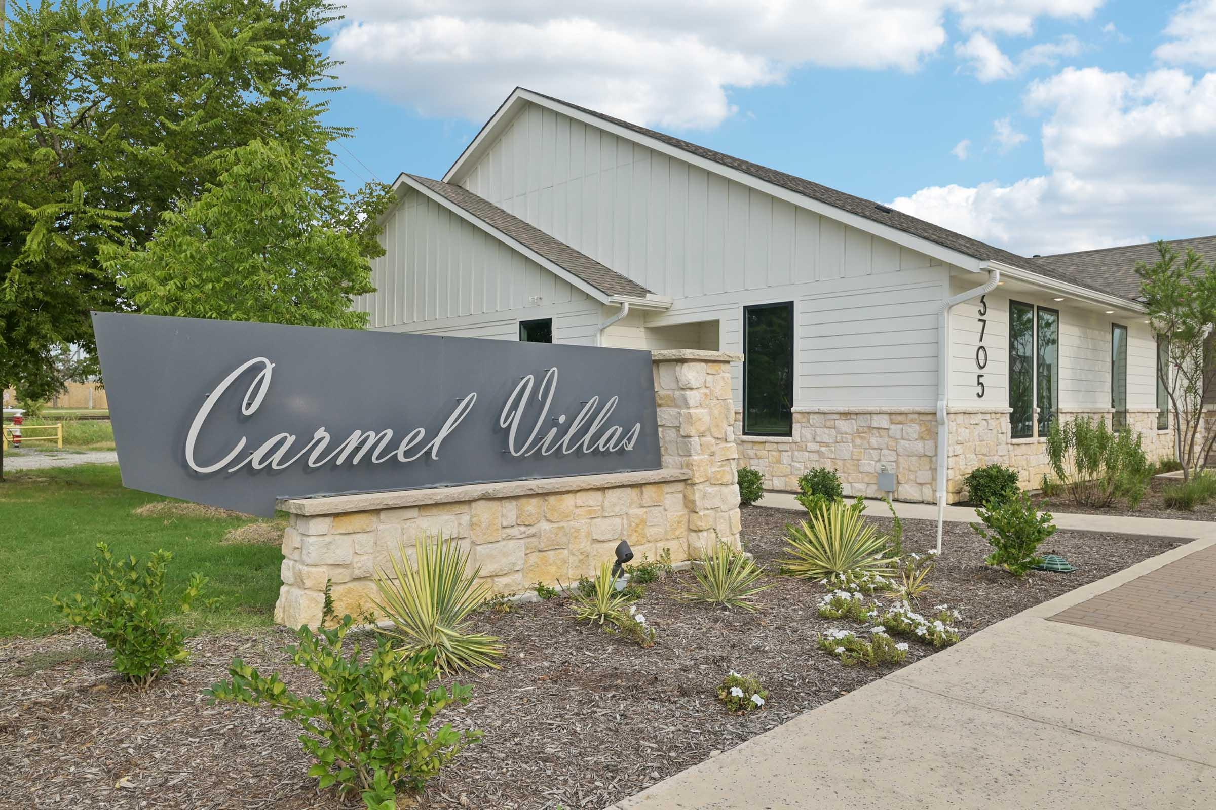 Sign for Carmel Villas in front of a single-story building, surrounded by landscaped bushes and grass. The building has a light-colored exterior with large windows, and the sky is partly cloudy.