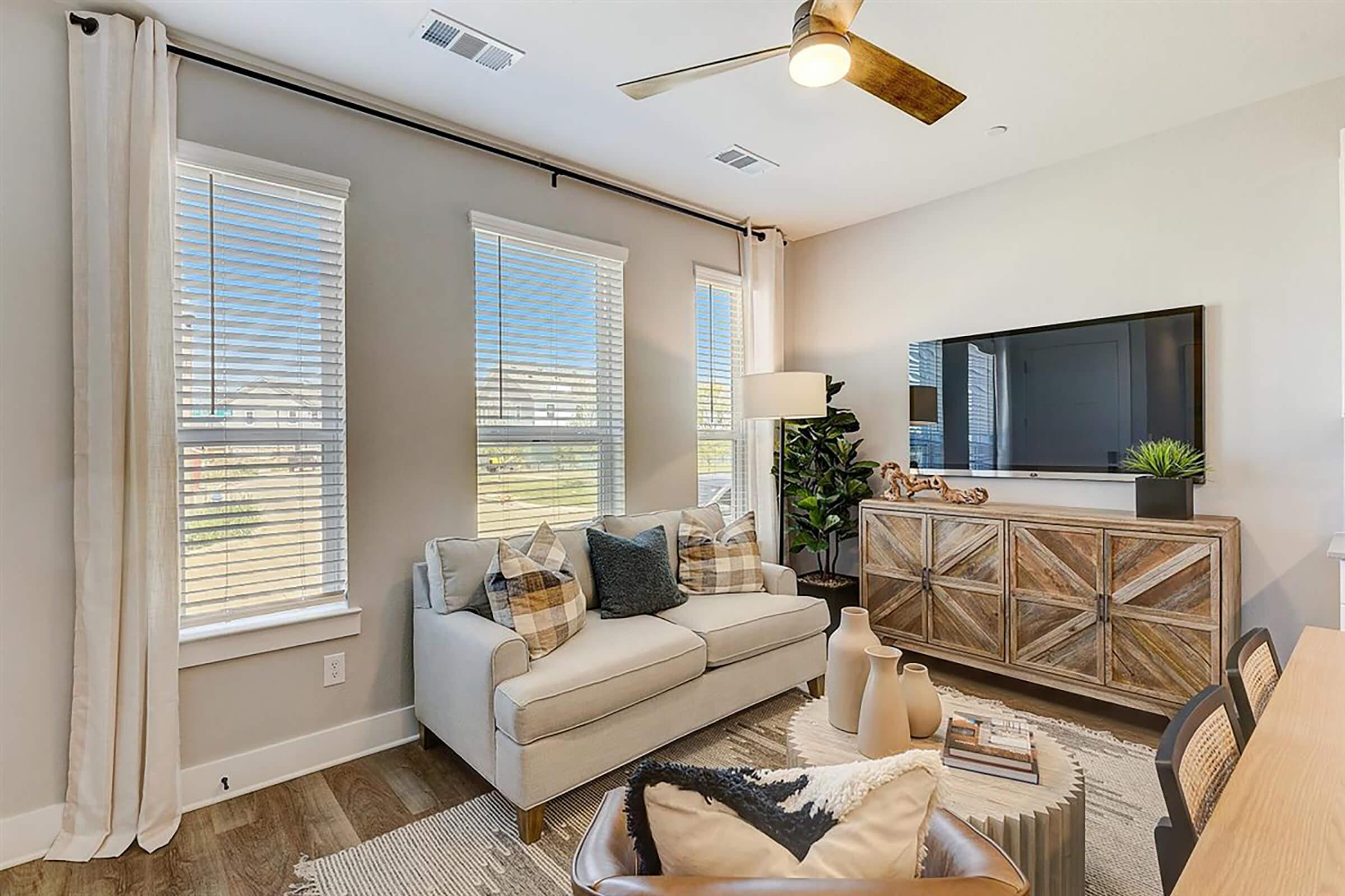 A modern living room featuring a light-colored sofa with decorative pillows, a wooden coffee table with a rug underneath, a TV mounted on the wall, and large windows with natural light. There are plants in the corner, adding a touch of greenery to the space, and a stylish sideboard against the wall.