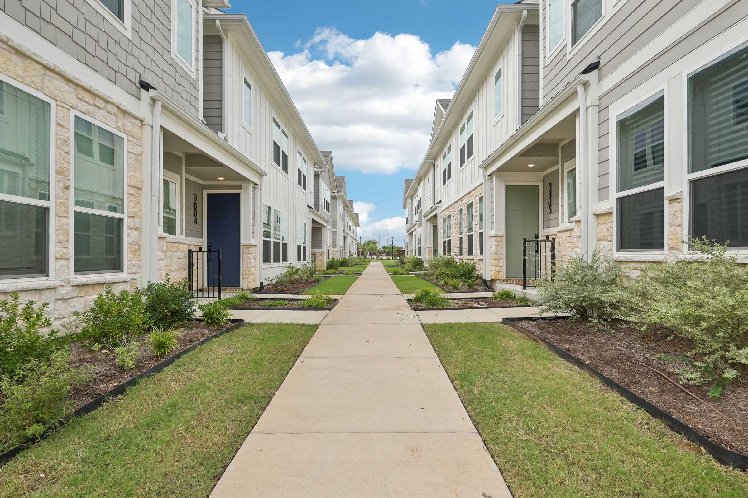 A wide, paved pathway runs between two rows of modern, two-story residential buildings. Each building has multiple windows and a front door, with neatly landscaped grass and plants lining the walkway. The sky above is partly cloudy, creating a bright and inviting atmosphere.