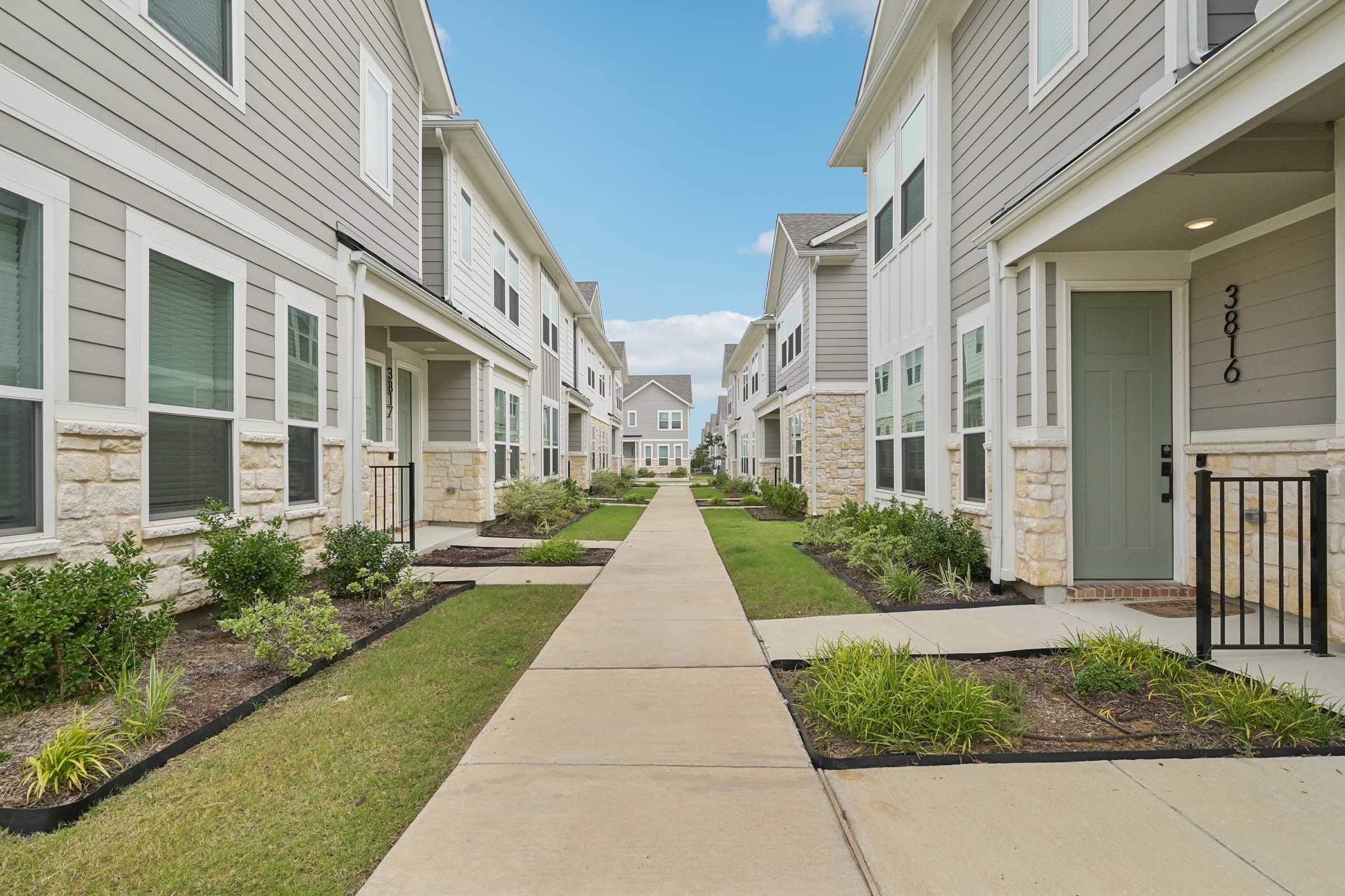 A view of a modern residential complex with two-story buildings on either side of a paved pathway. Lush green grass and tidy landscaping line the walkway, leading towards more buildings in the distance under a blue sky. The architecture features a mix of stone and siding.