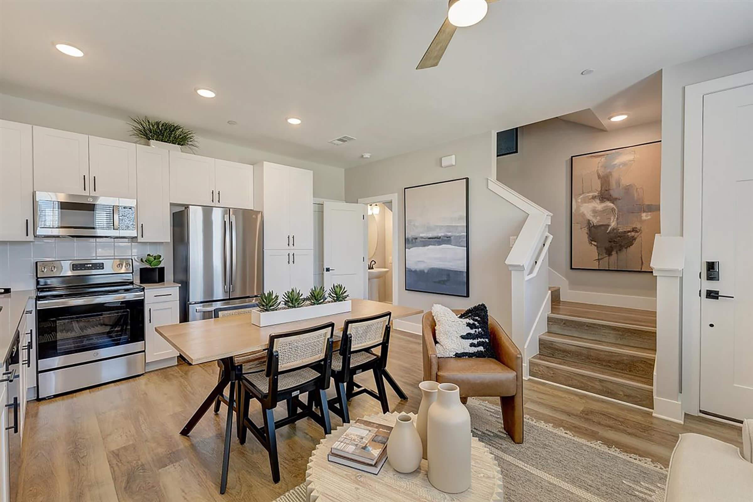 Modern open-concept kitchen and living area featuring stainless steel appliances, a wooden dining table with black chairs, and a beige leather sofa. Stairs lead to an upper level, while a large abstract painting adorns the wall. Natural light fills the space, creating a warm and inviting atmosphere.