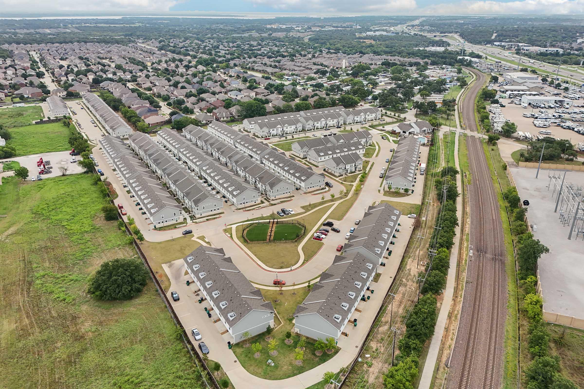 Aerial view of a suburban neighborhood featuring rows of similar houses, winding pathways, and green spaces. Adjacent to the neighborhood are railroad tracks and a highway, with additional residential areas visible in the background. The scene captures a mix of urban and natural elements.