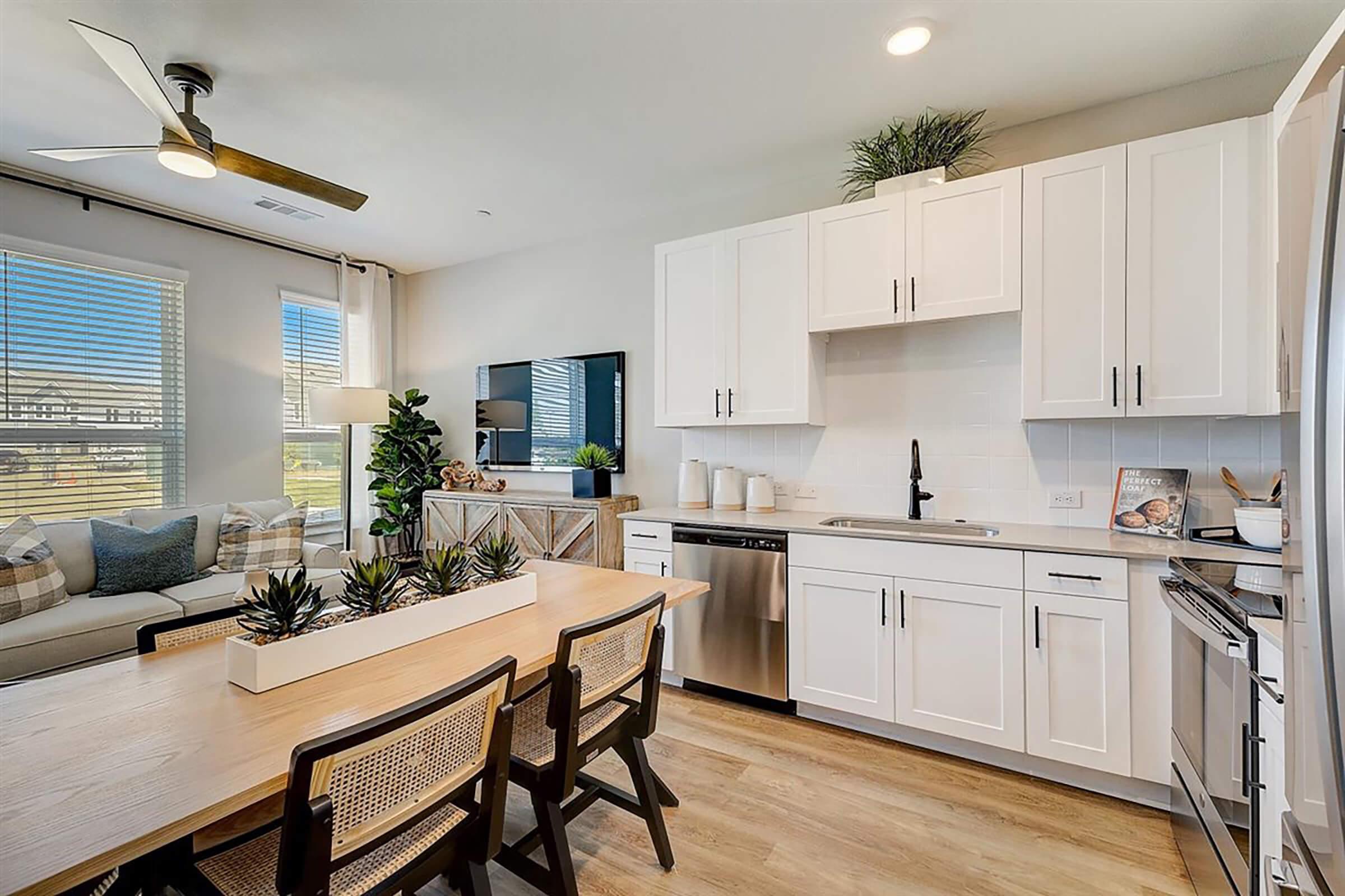 Bright and modern kitchen featuring white cabinetry, stainless steel appliances, and a dining area with a wooden table and woven chairs. A large window provides natural light, and decorative plants add greenery to the space. The living area is visible in the background, adding a cozy touch.