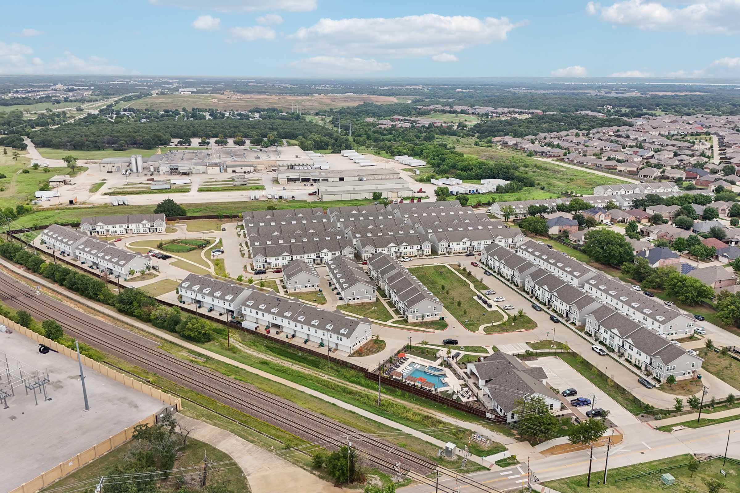 Aerial view of a residential community featuring multiple buildings arranged in a circular layout, a swimming pool in the center, greenery surrounding the area, and nearby industrial structures in the background. The landscape includes a railway line and expansive fields.